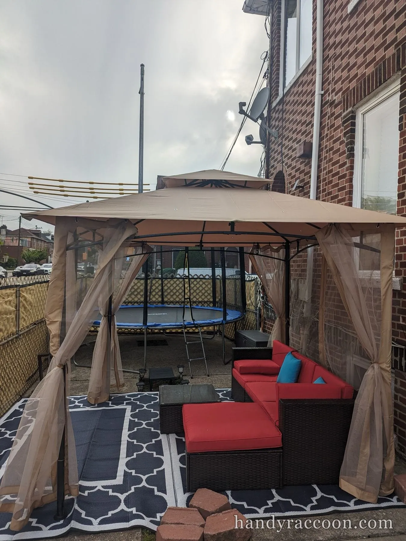 Beige canopy gazebo with mesh sides over a red wicker sectional sofa on a patterned rug in a backyard.