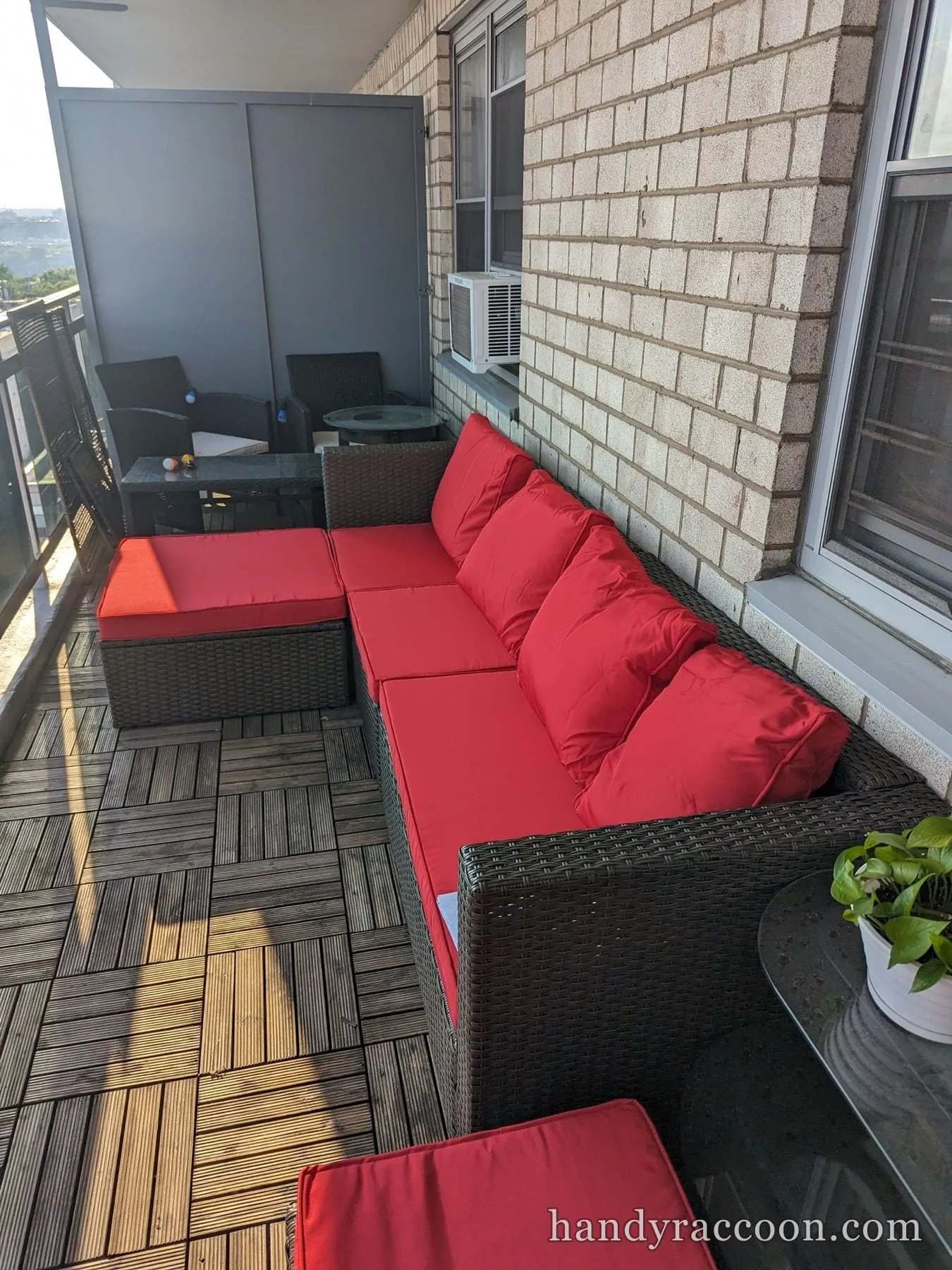 Dark wicker sectional sofa with red cushions assembled along a brick wall on a wood-tiled balcony.