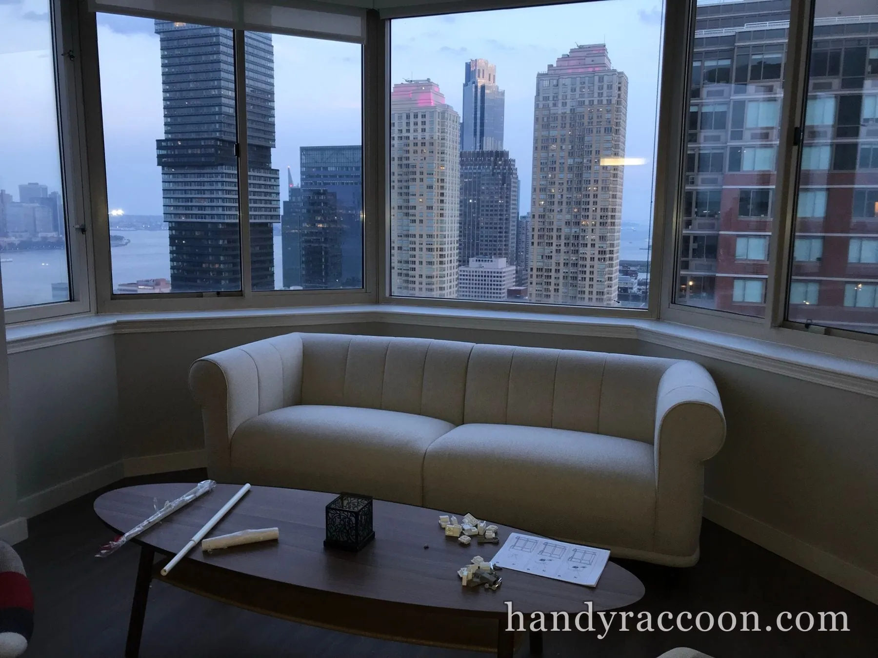 Cream channel-tufted sofa assembled in front of corner windows overlooking a city skyline at dusk