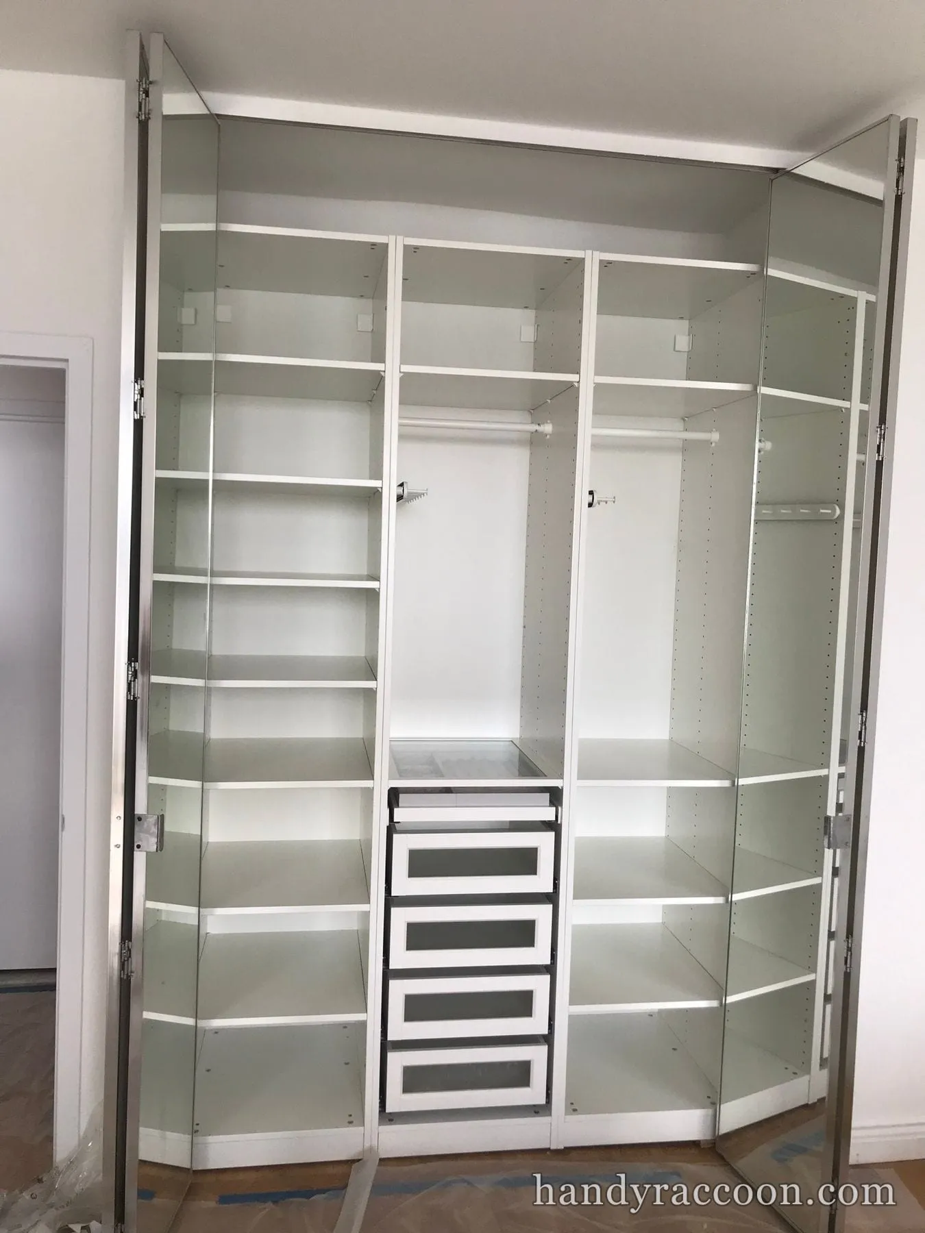 White corner wardrobe with mirrored doors open showing symmetrical shelves, rails and a drawer tower
