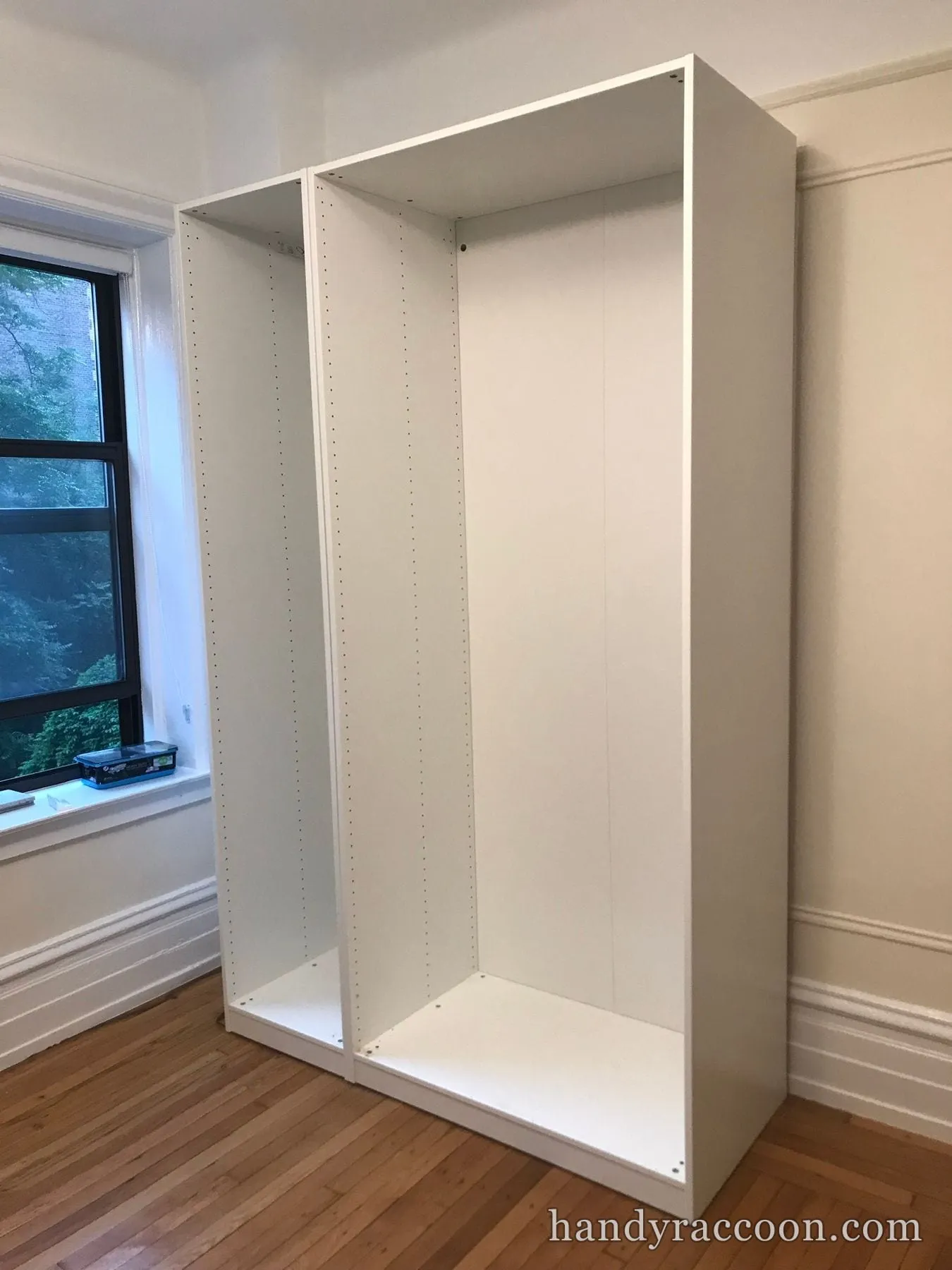 Two white tall wardrobe carcass frames assembled and standing by a bedroom window.