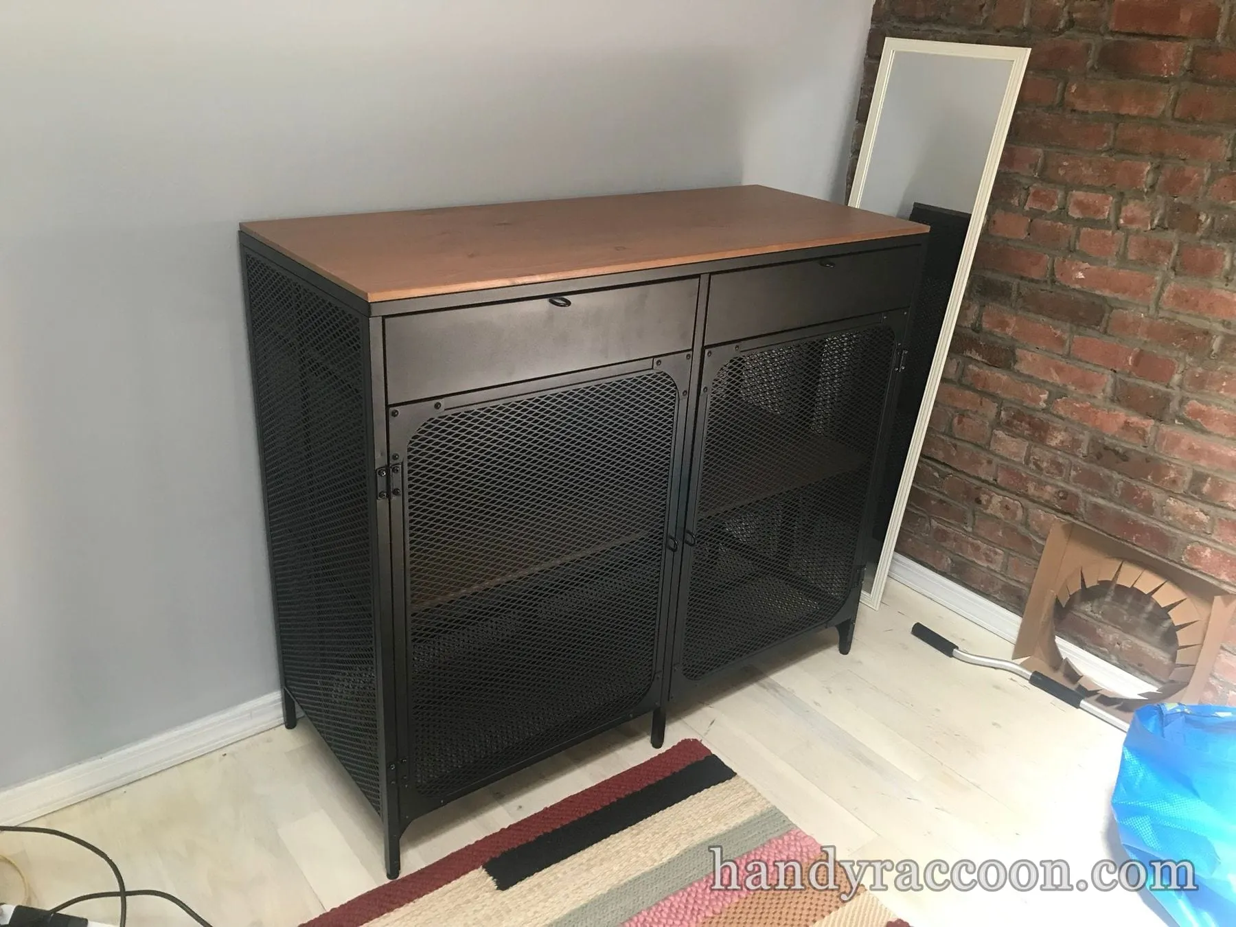 Assembled black metal mesh storage cabinet with two drawers and a light wood top near an exposed brick wall.
