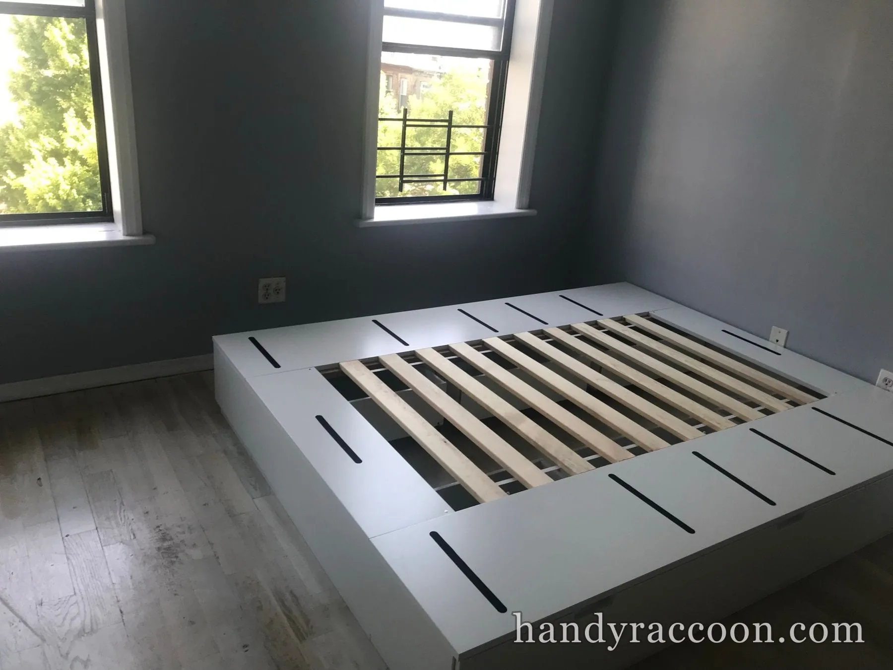 Assembled white platform bed base with light wood slats in a gray-walled bedroom with two windows.