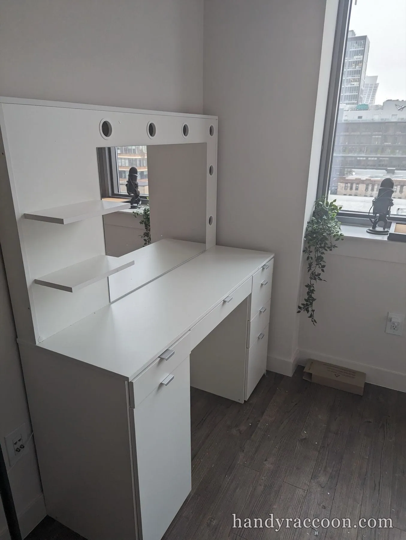 White makeup vanity with a light-bulb mirror, side shelves and drawers assembled in a corner by a window.