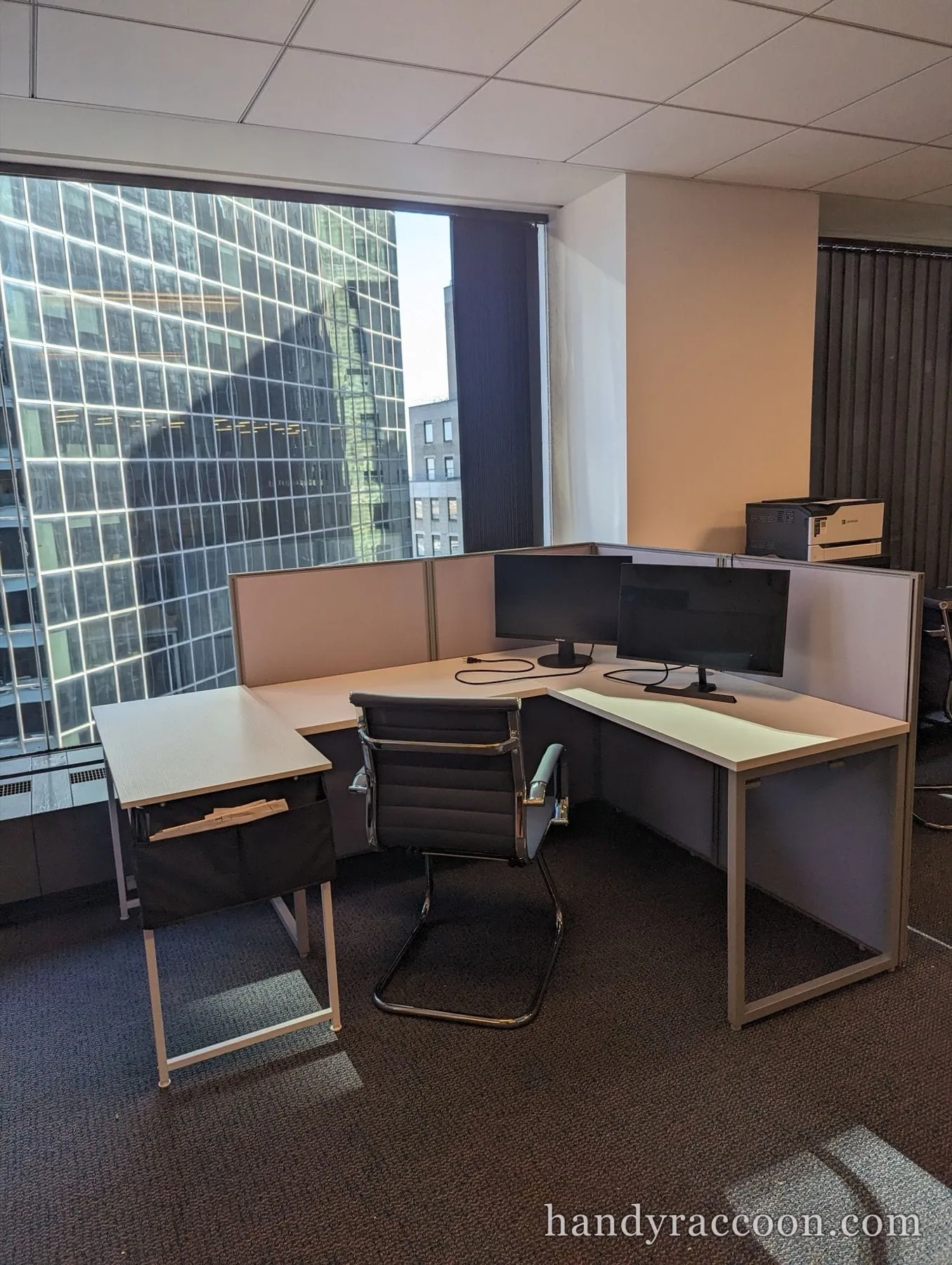 Assembled L-shaped office desk with two monitors, an office chair and a printer by an office window.