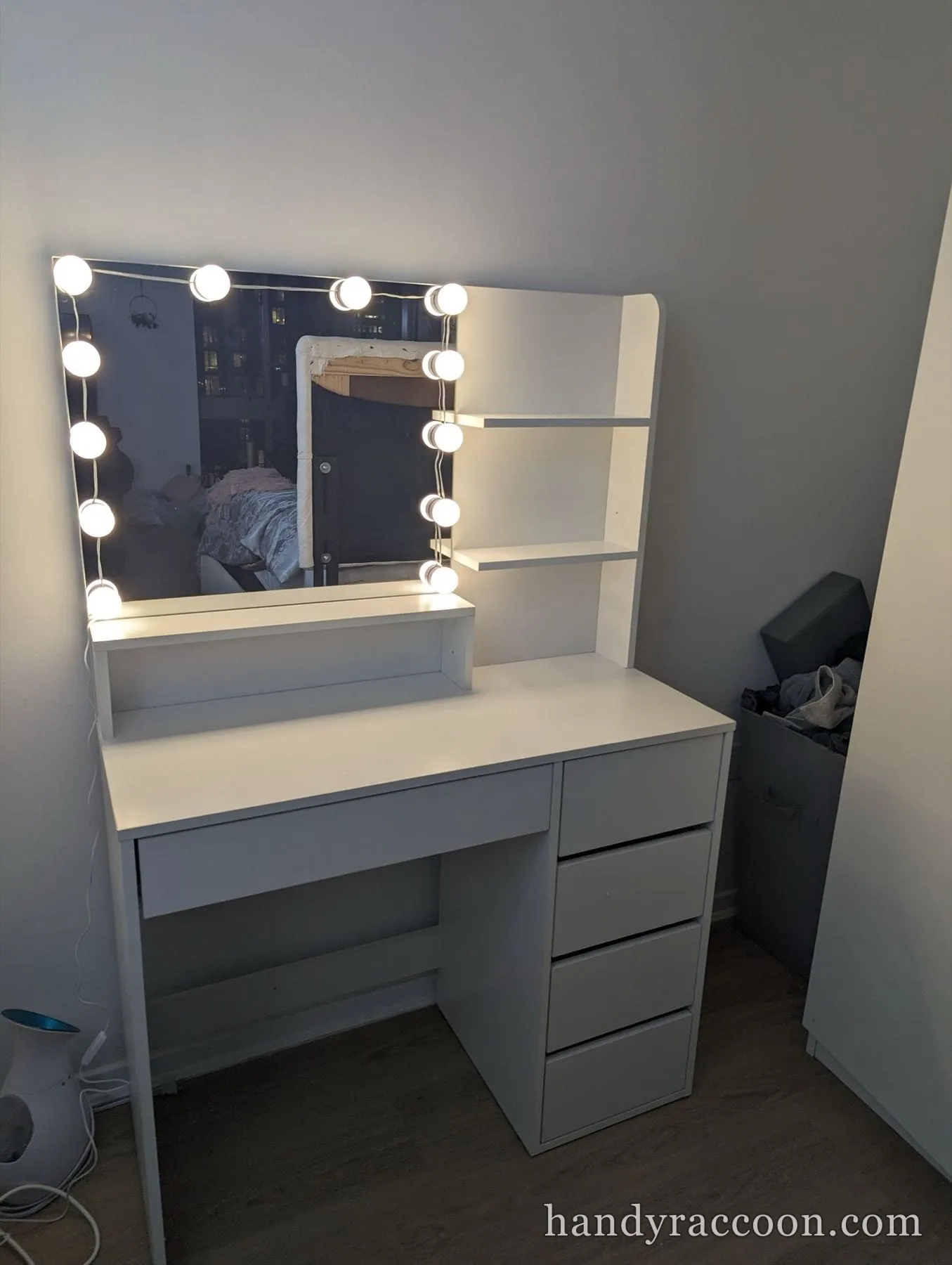 White makeup vanity with an illuminated bulb-framed mirror, side shelf and four-drawer tower in a bedroom.