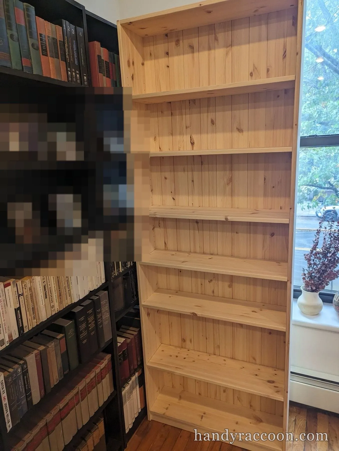 Tall unfinished pine bookcase with empty shelves assembled beside a black bookshelf full of books by a window.