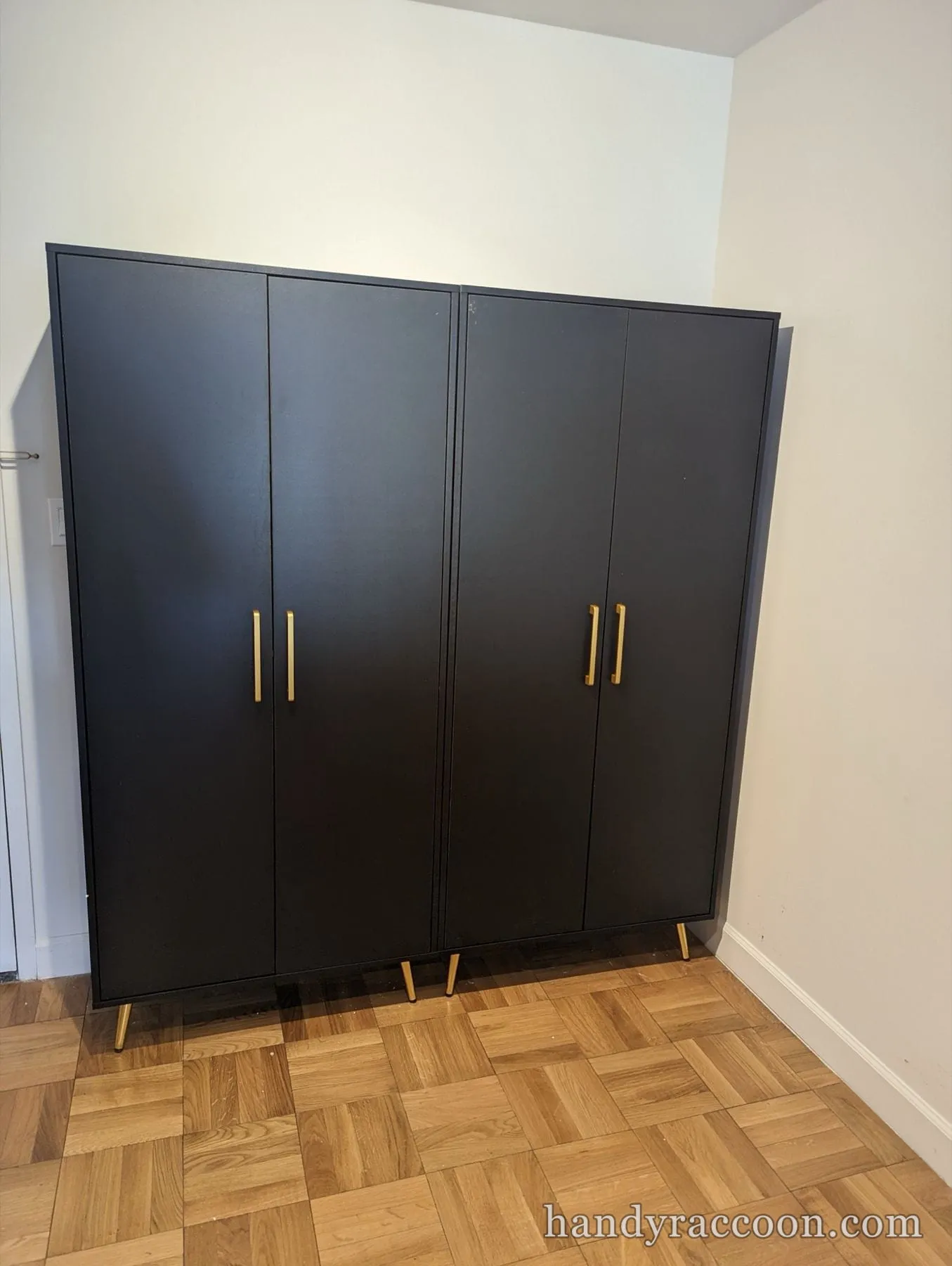 Two black double-door wardrobe cabinets with brass handles and legs on parquet flooring.