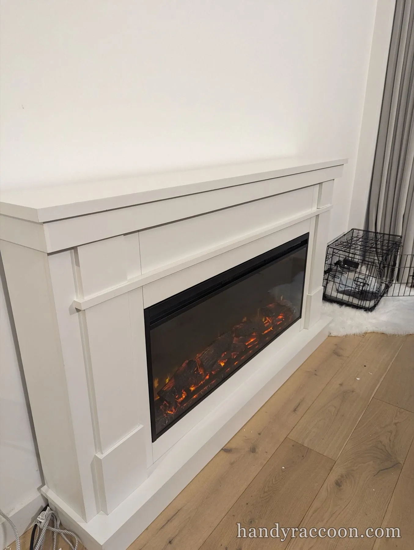 White fireplace mantel with a lit electric flame insert against a white wall on wood floor.