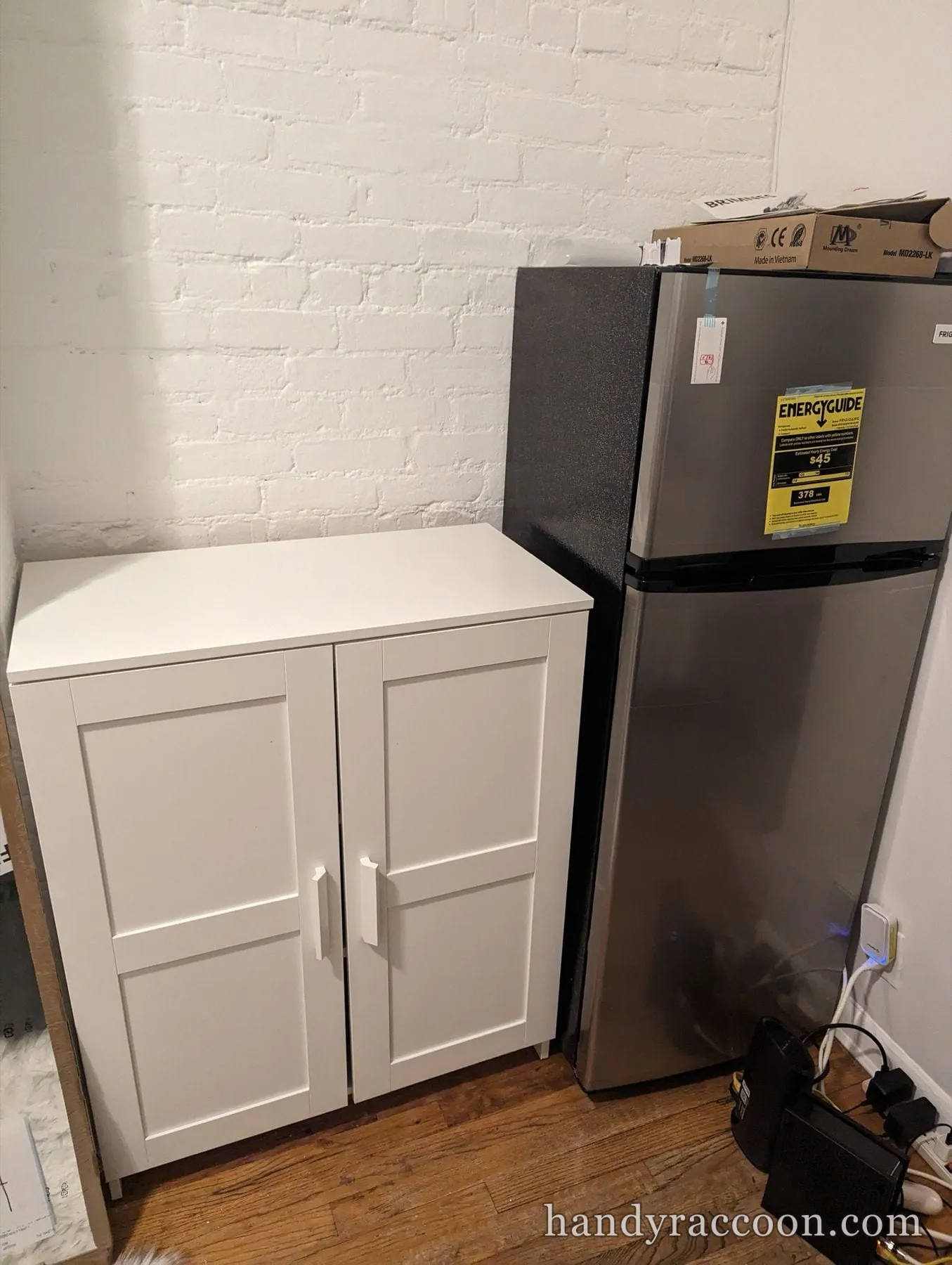White two-door storage cabinet next to a stainless refrigerator on a wood floor.