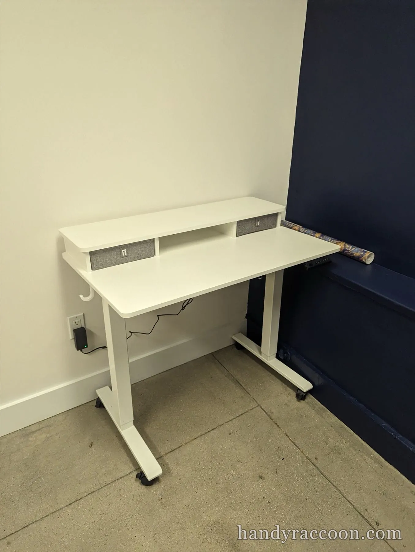 White standing desk on wheels with a two-drawer shelf riser against a white wall.