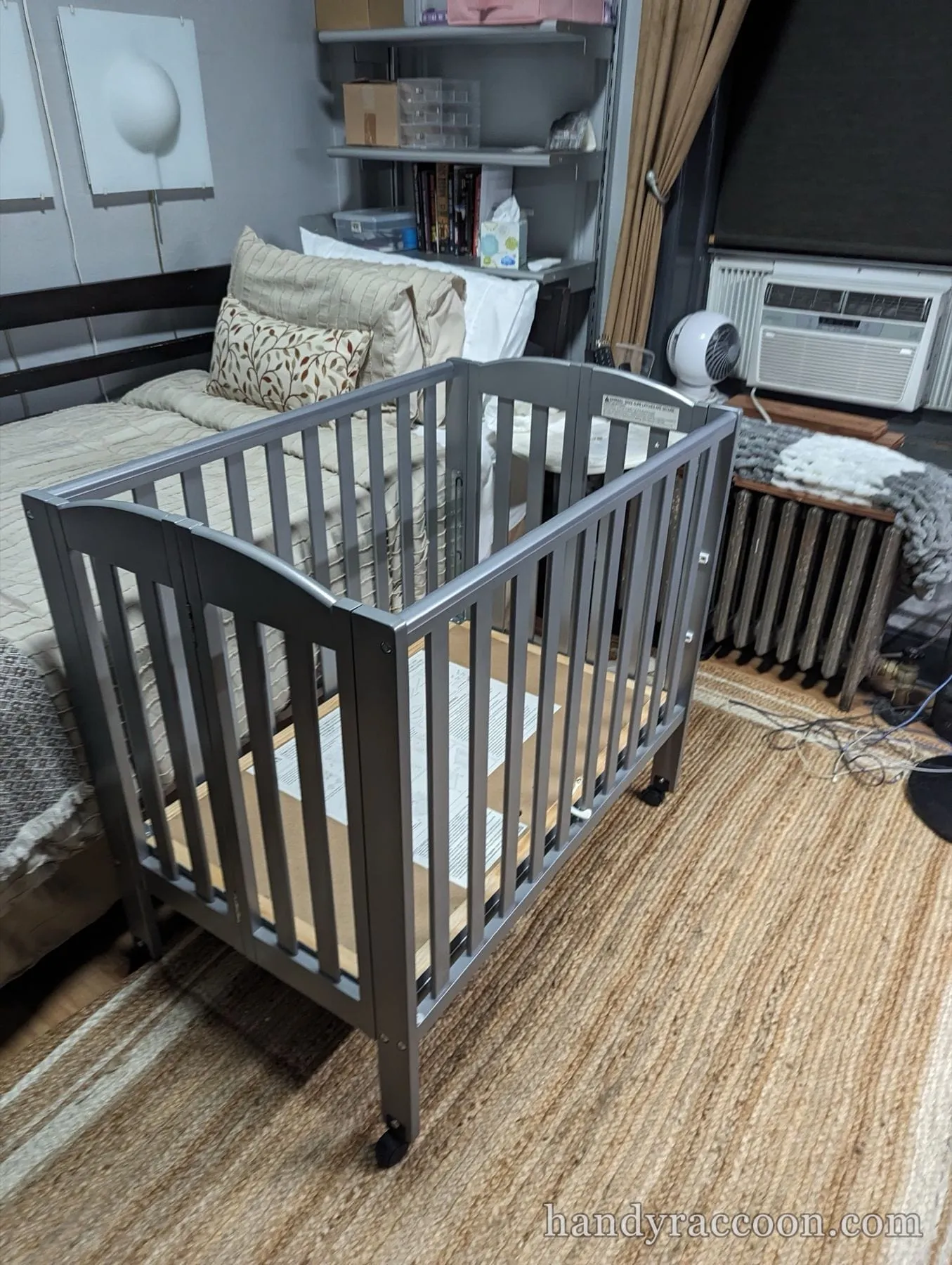 Assembled grey slatted mini crib on wheels beside a bed on a woven rug.