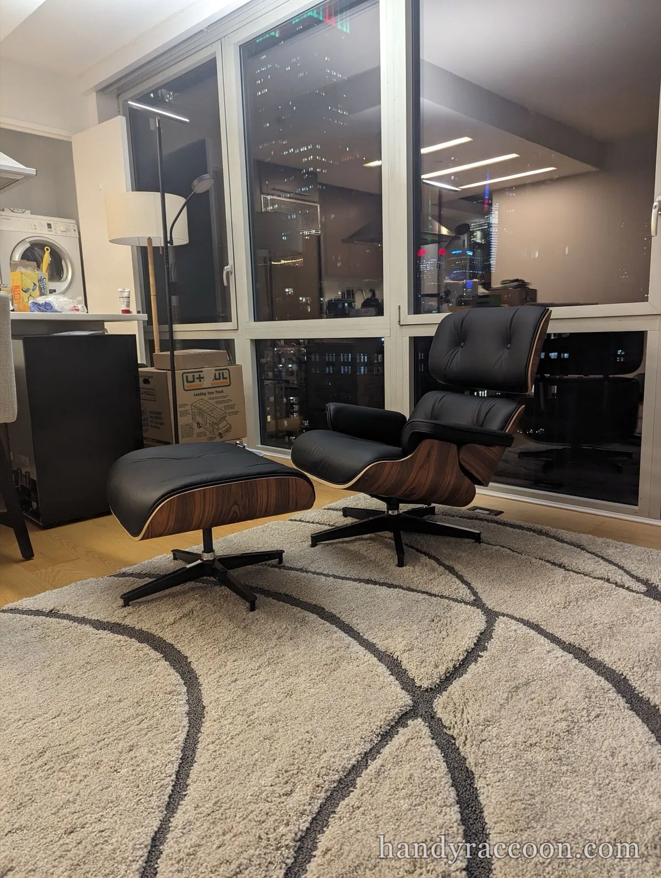 Black leather lounge chair and ottoman on a patterned rug in front of tall windows at night.
