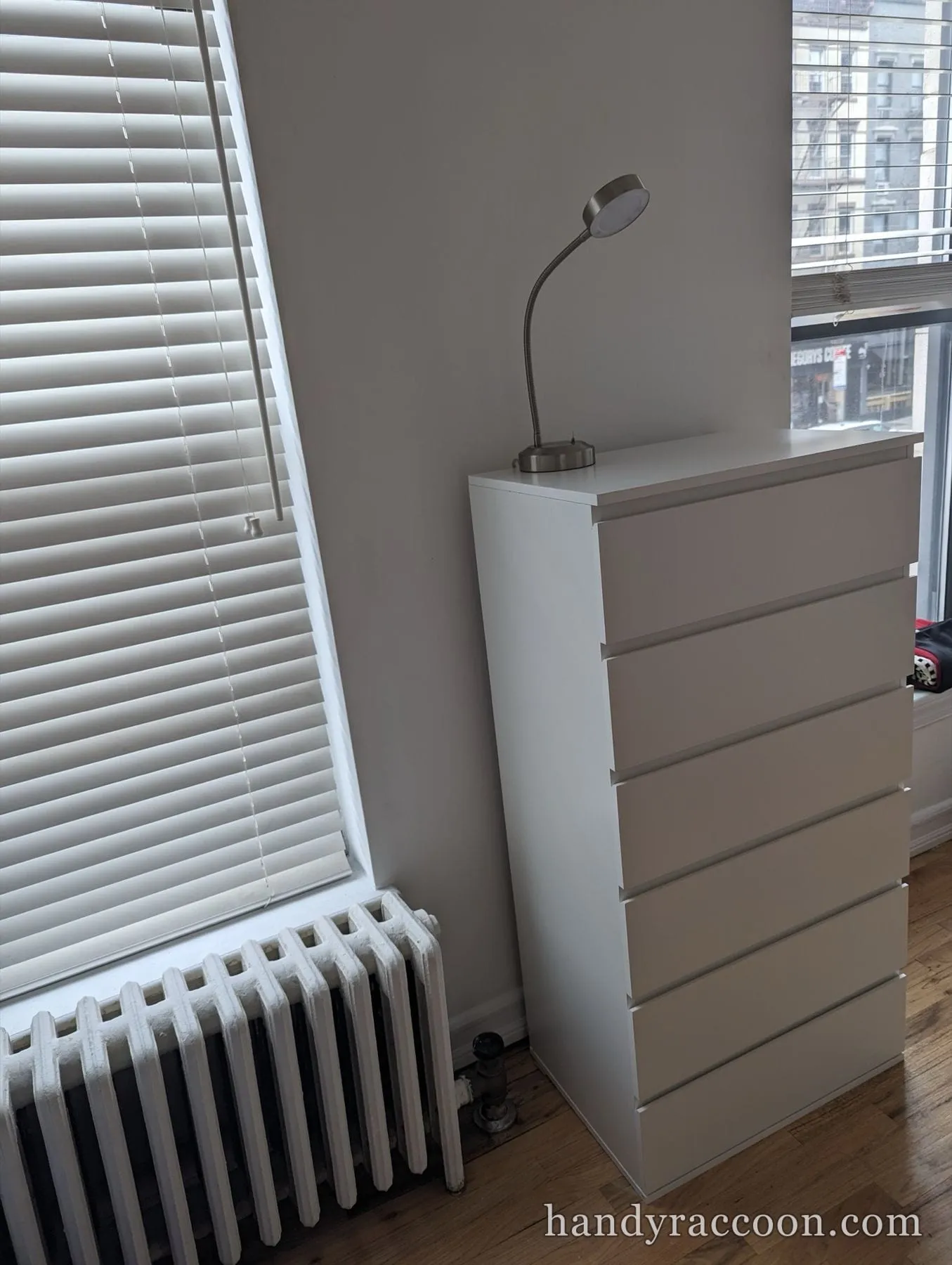 Tall white six-drawer dresser next to a window with blinds and a painted radiator.