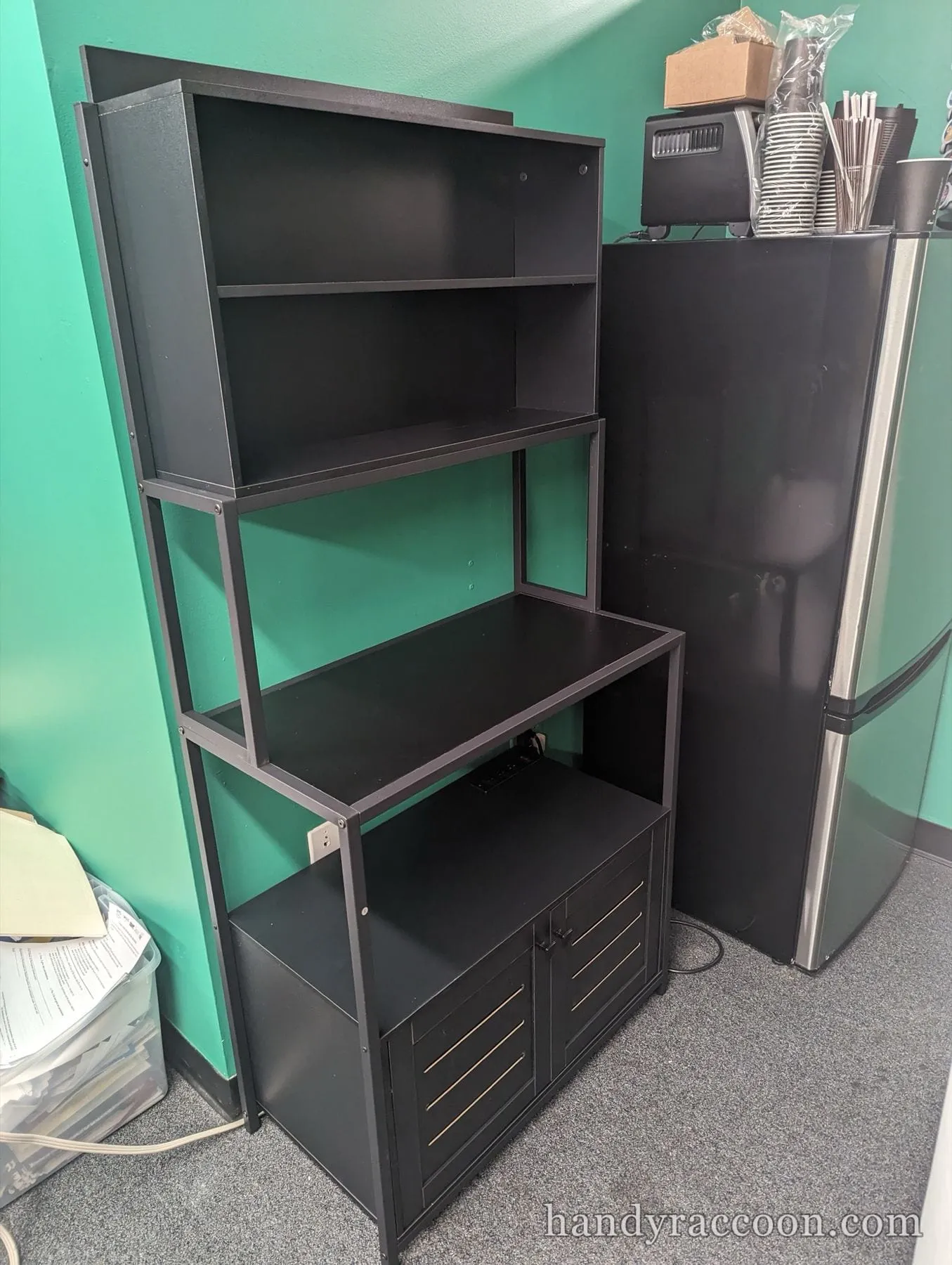 Tall black bakers rack style shelving unit with a lower cabinet against a green wall.