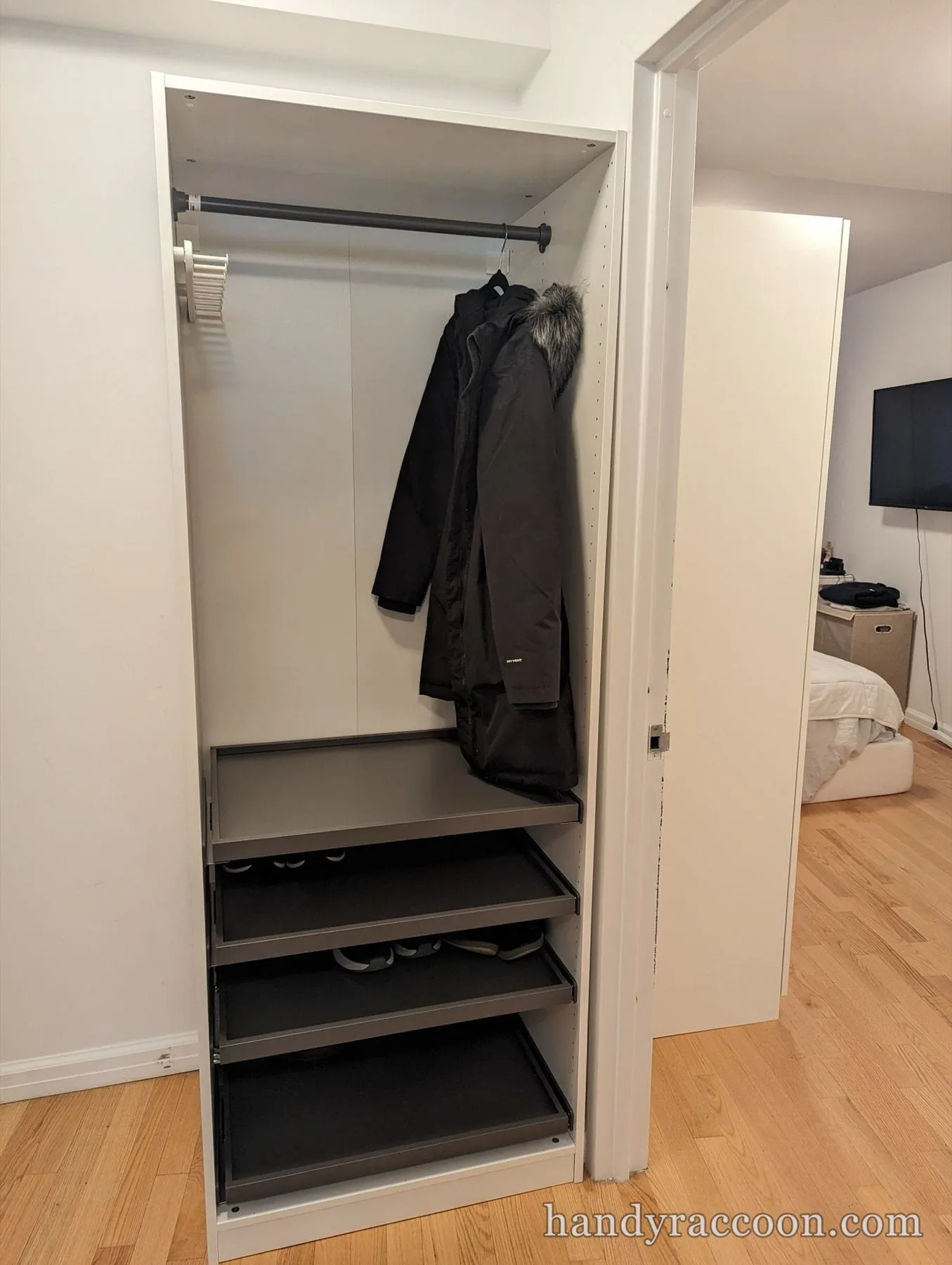Open white wardrobe with a hanging coat, closet rod and pull-out shelves on a wood floor.