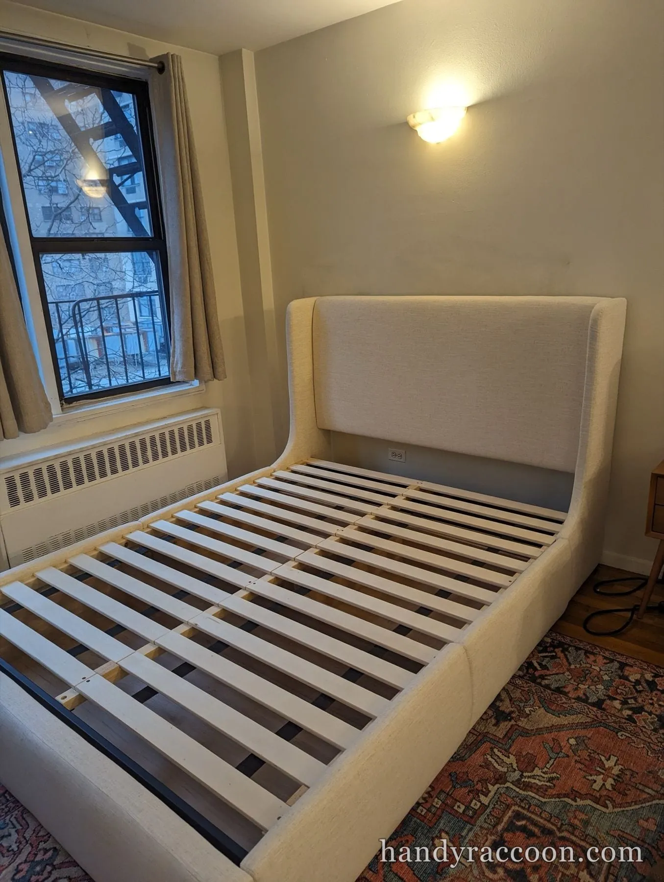 Cream upholstered wingback bed frame with wood slats on a patterned rug near a window.