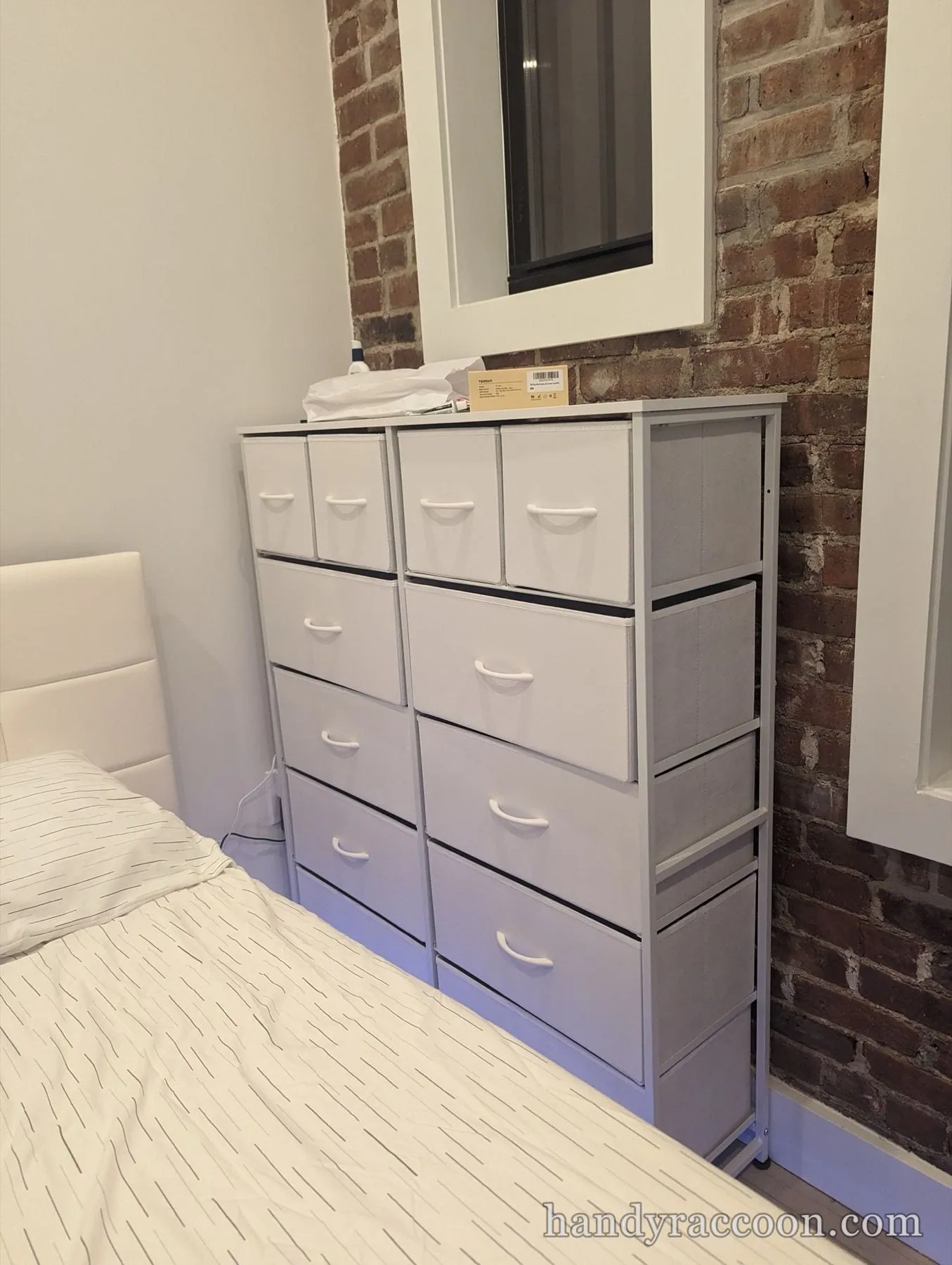 Tall white fabric drawer dresser next to an upholstered bed and exposed brick wall.