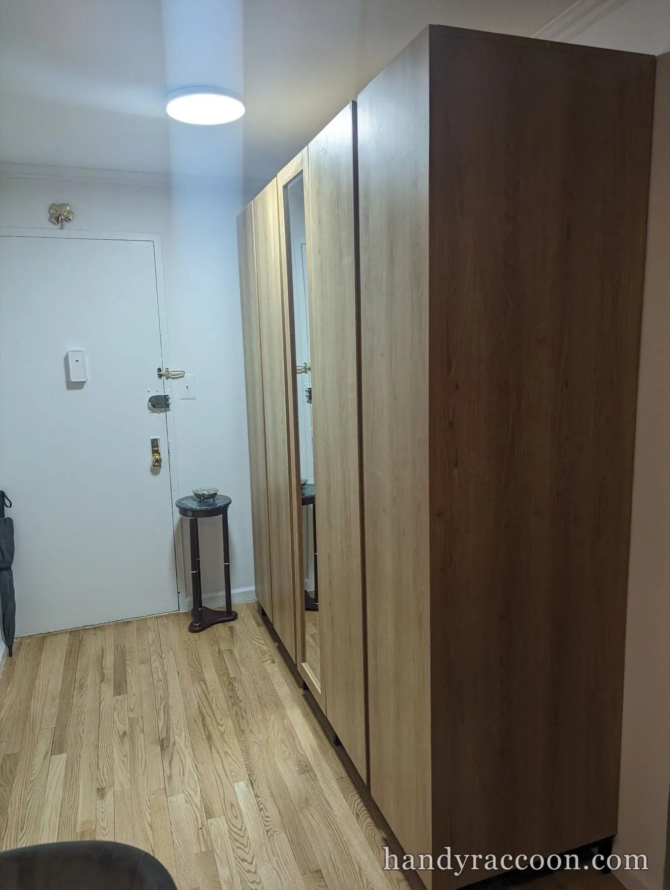 Tall wood-finish wardrobe with a mirrored door standing along a hallway wall near an apartment door.