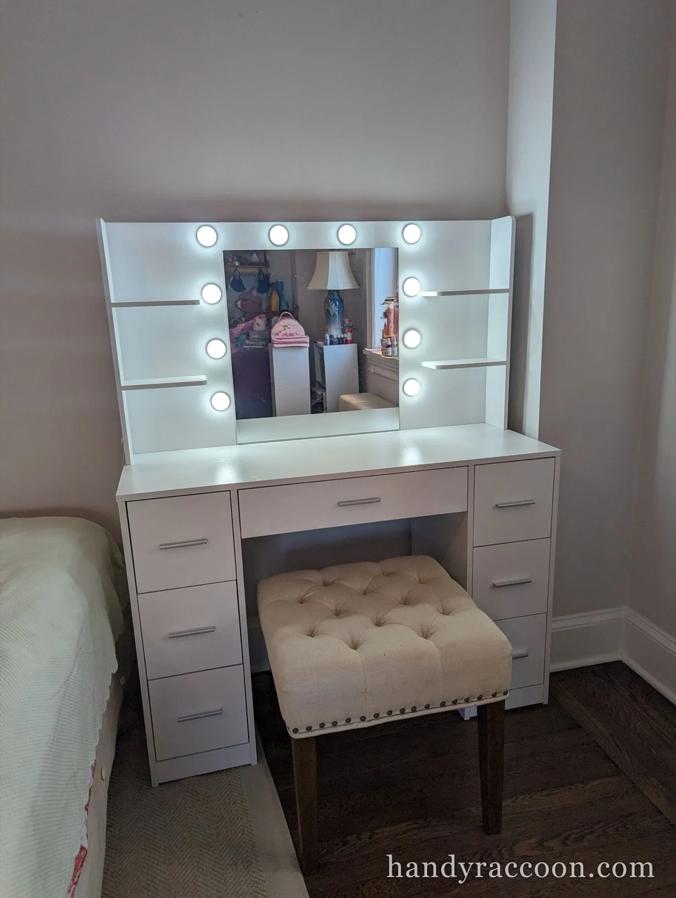 White makeup vanity desk with a light-bulb mirror and side drawers, an upholstered stool tucked beneath.