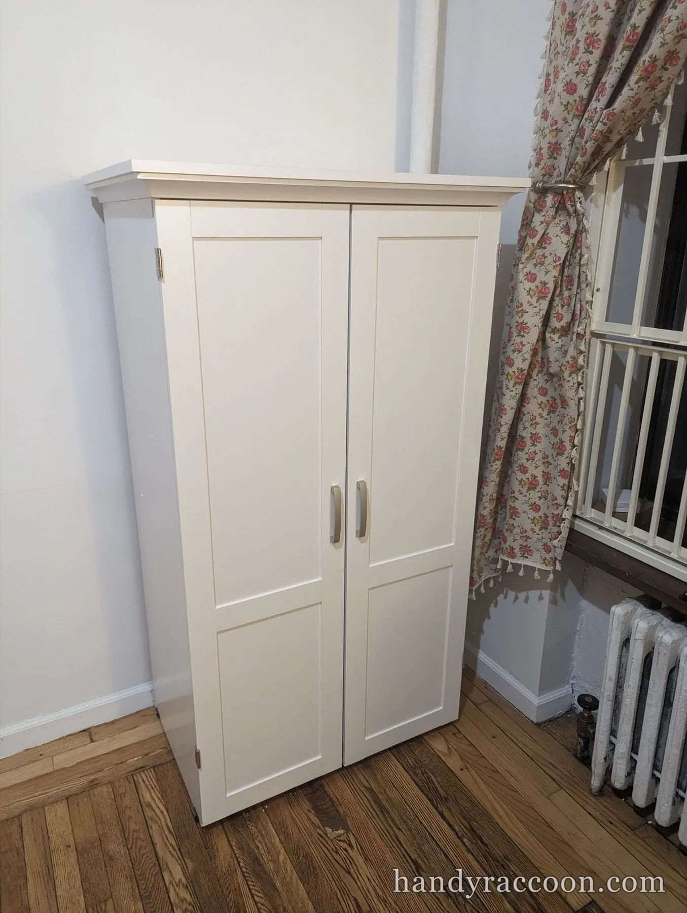 White two-door shaker cabinet assembled in a corner beside a floral curtain and radiator.