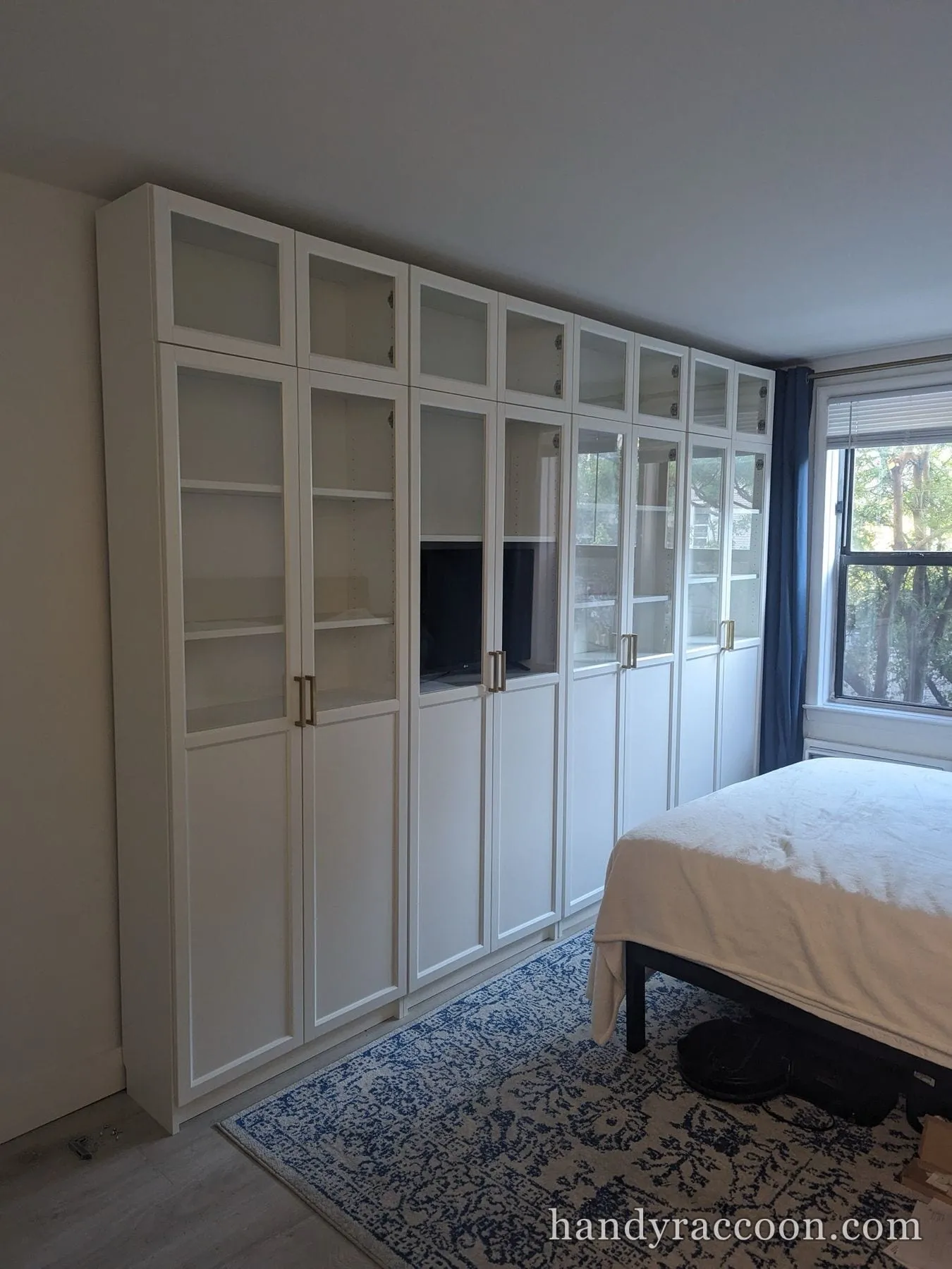 Tall white cabinet and bookcase system with glass doors assembled along a bedroom wall.