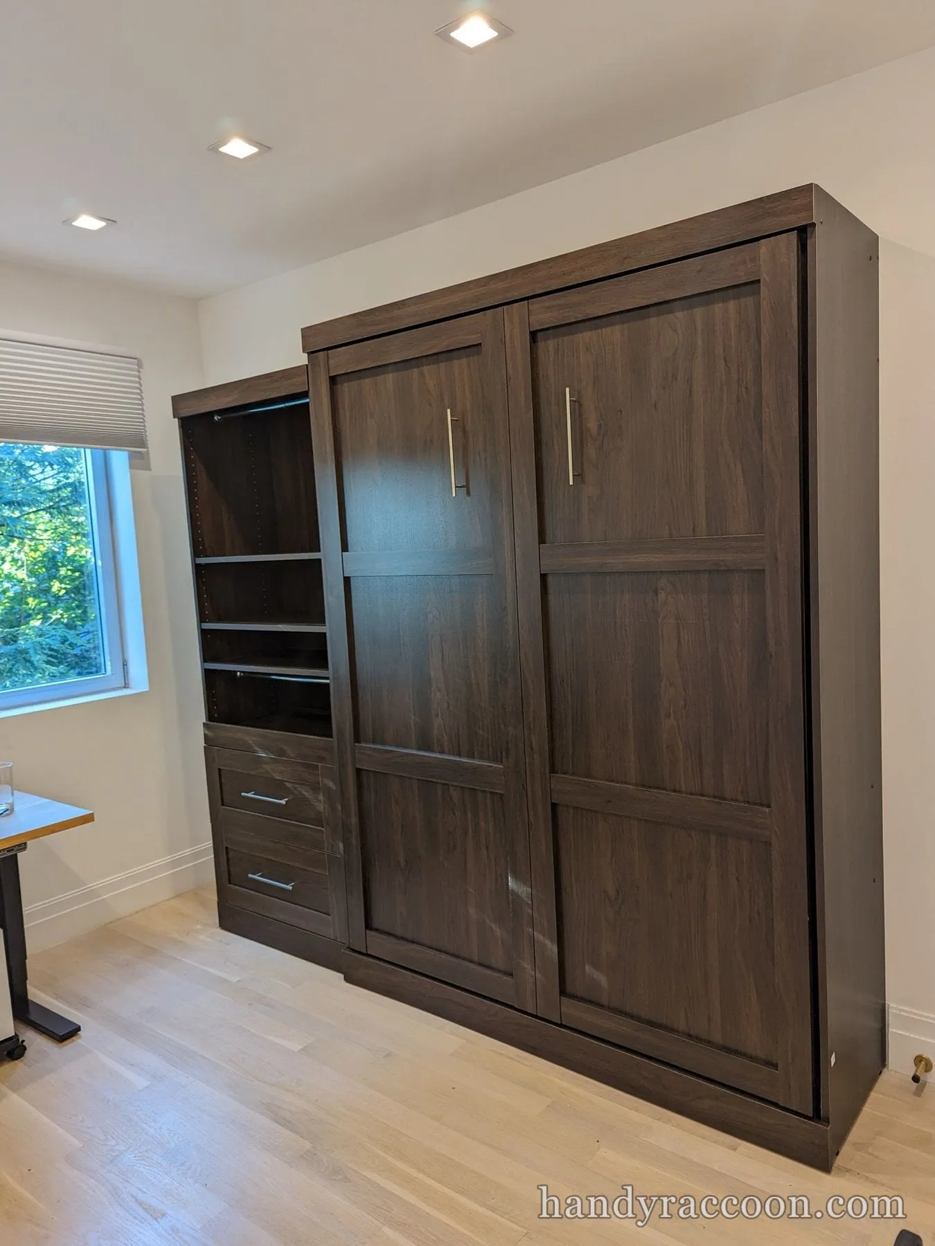 Closed dark wood-tone Murphy wall bed cabinet next to a matching shelving and drawer unit in a bright room.