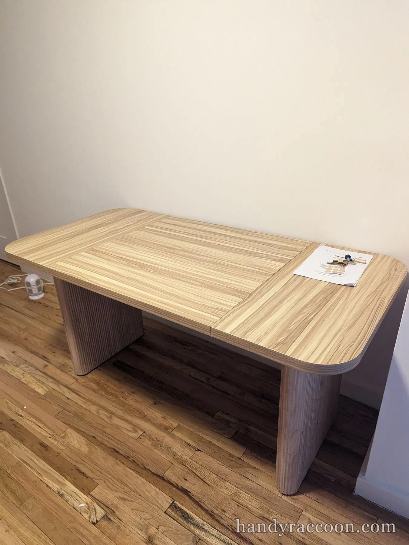 Light wood coffee table with fluted pedestal legs on a hardwood floor, assembly sheet on top.