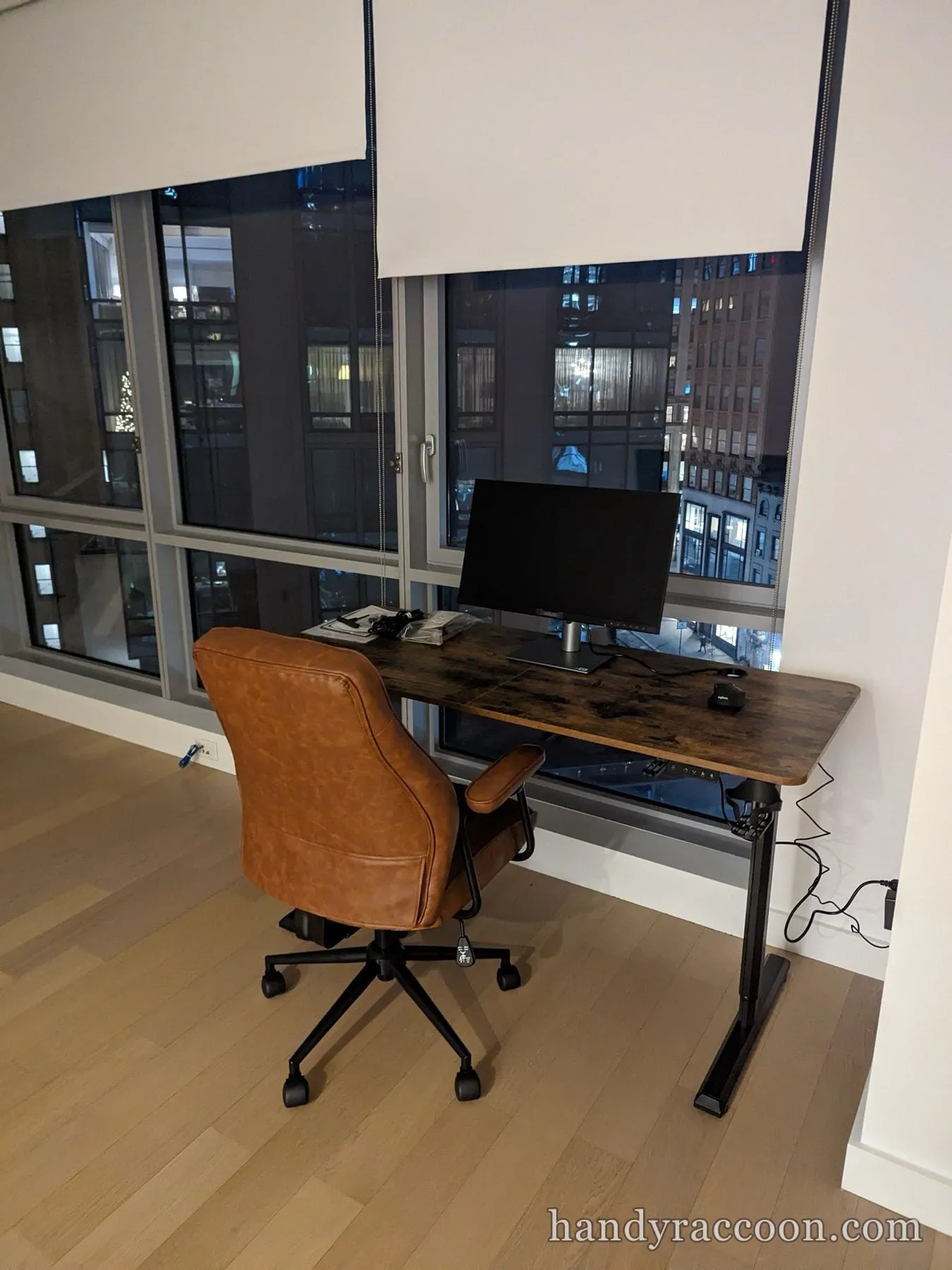 Rustic-top height-adjustable desk with a monitor and brown leather office chair by a night window.