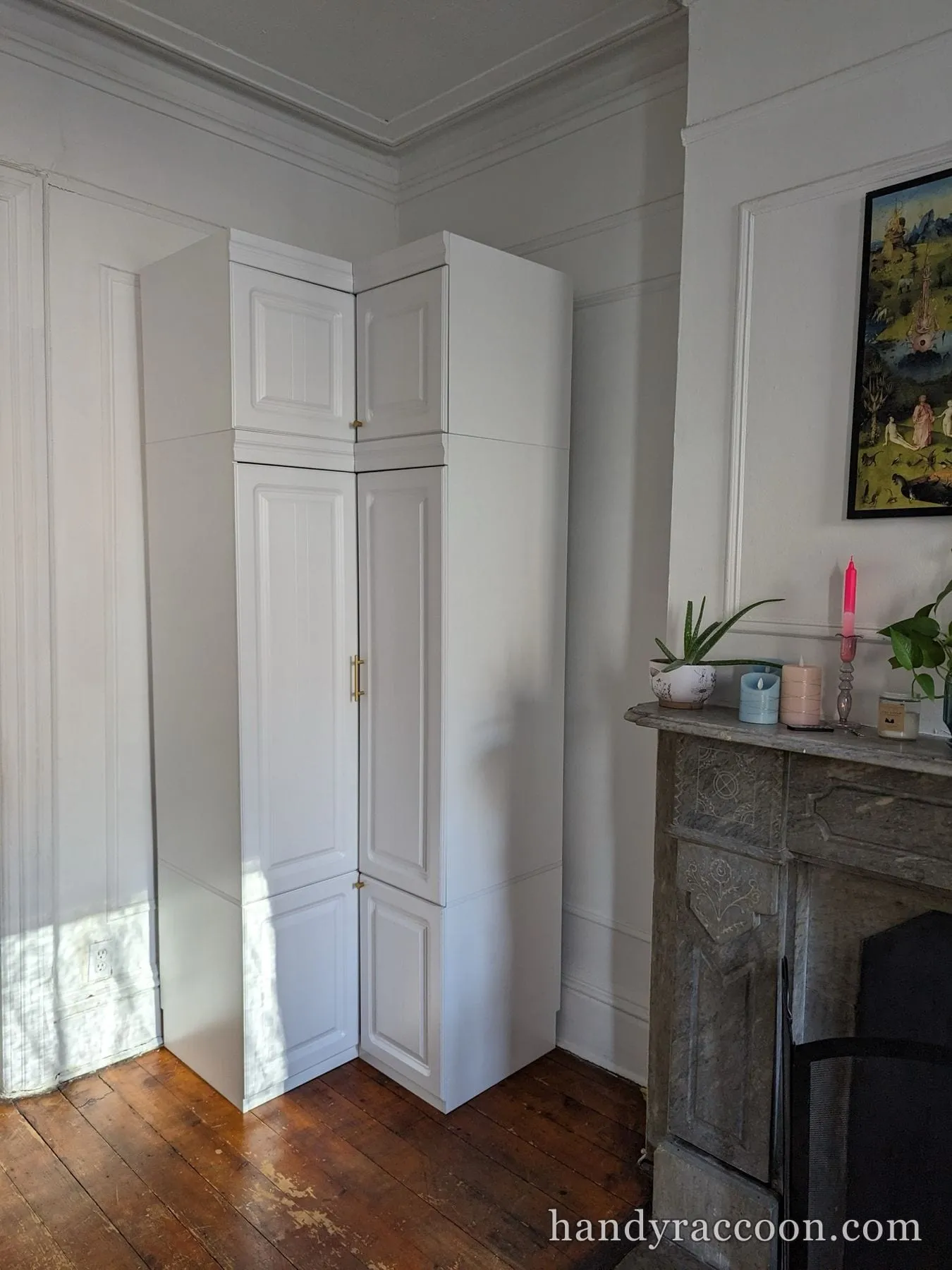 Two stacked white corner wardrobe cabinets with brass handles beside a marble fireplace.