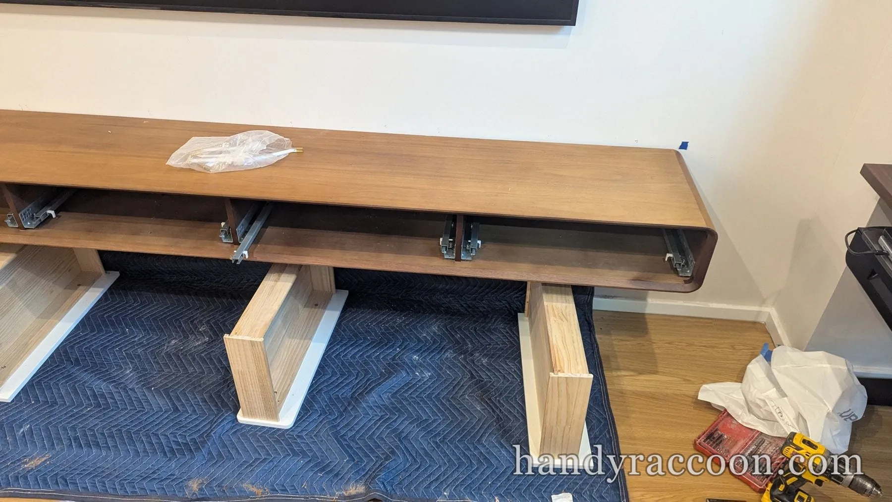 Long walnut media console on a moving blanket with drawer slides exposed and a drill and bit set nearby.