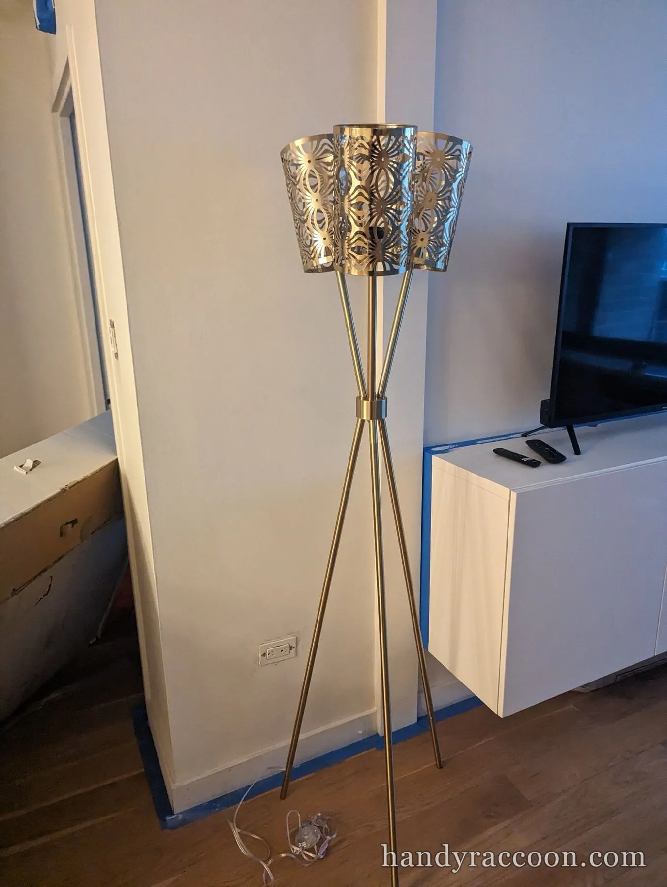 Gold tripod floor lamp with a cut-metal drum shade standing beside a media cabinet.