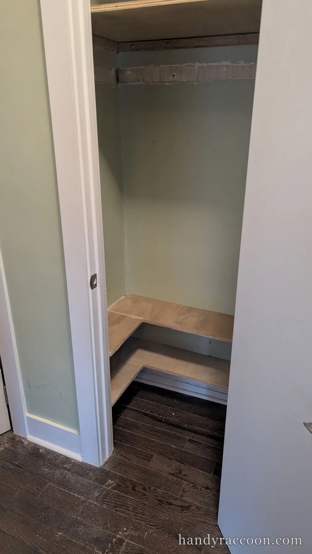 Interior of a closet with a newly built L-shaped plywood corner shelf against sage green walls.