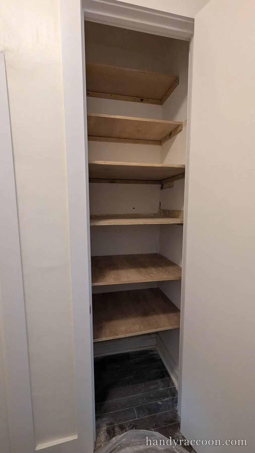 Tall narrow closet fitted with five stacked plywood shelves and white painted walls.