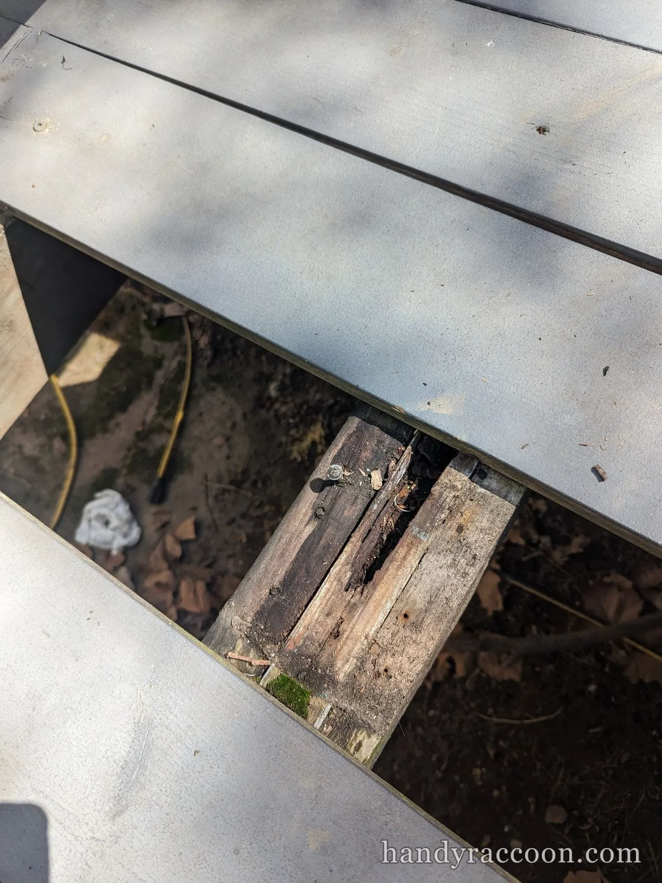 Looking down through a gap in gray deck boards at rotted wood framing and dirt below.