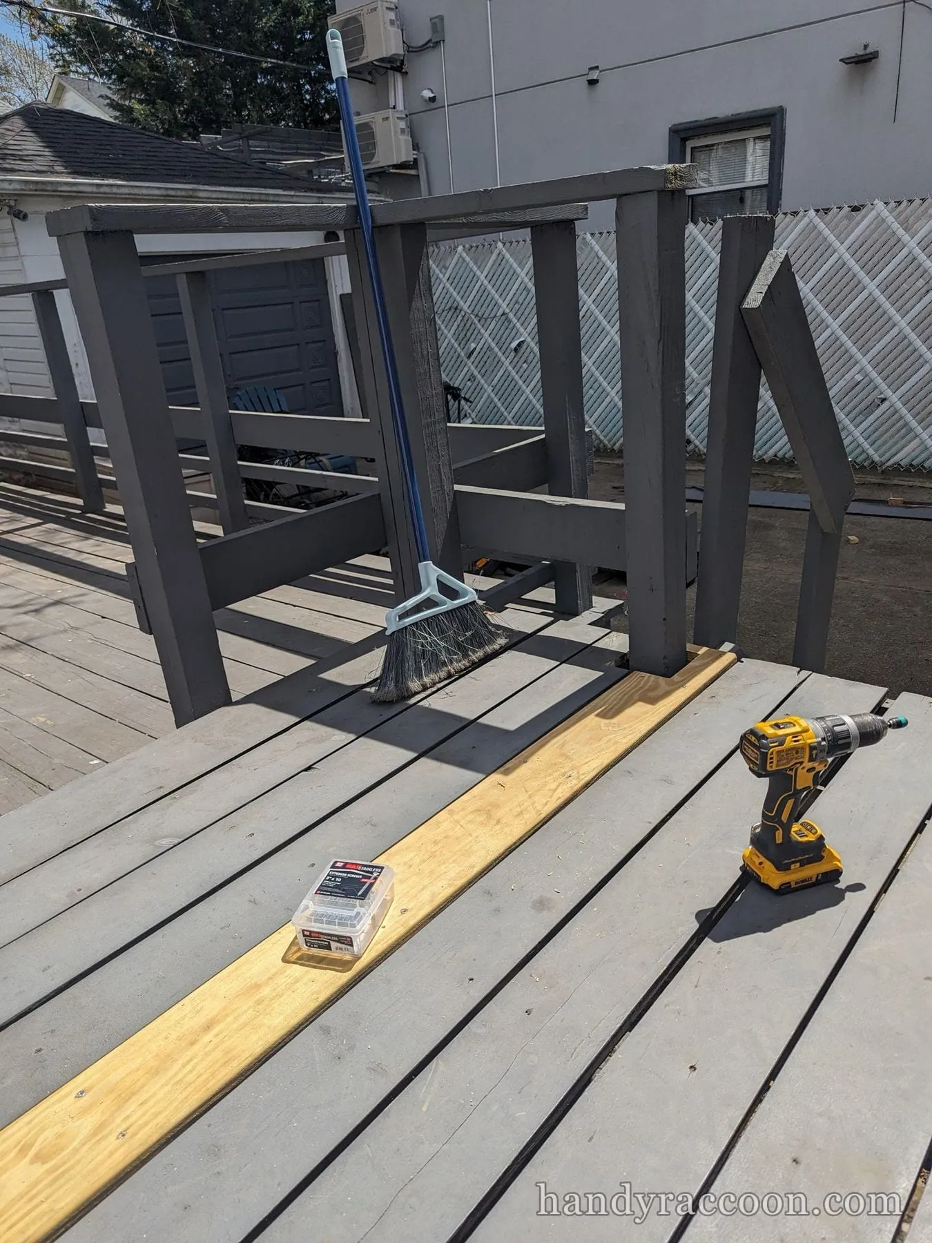 New wood deck board laid on a gray deck beside a cordless drill, screw box, and broom.