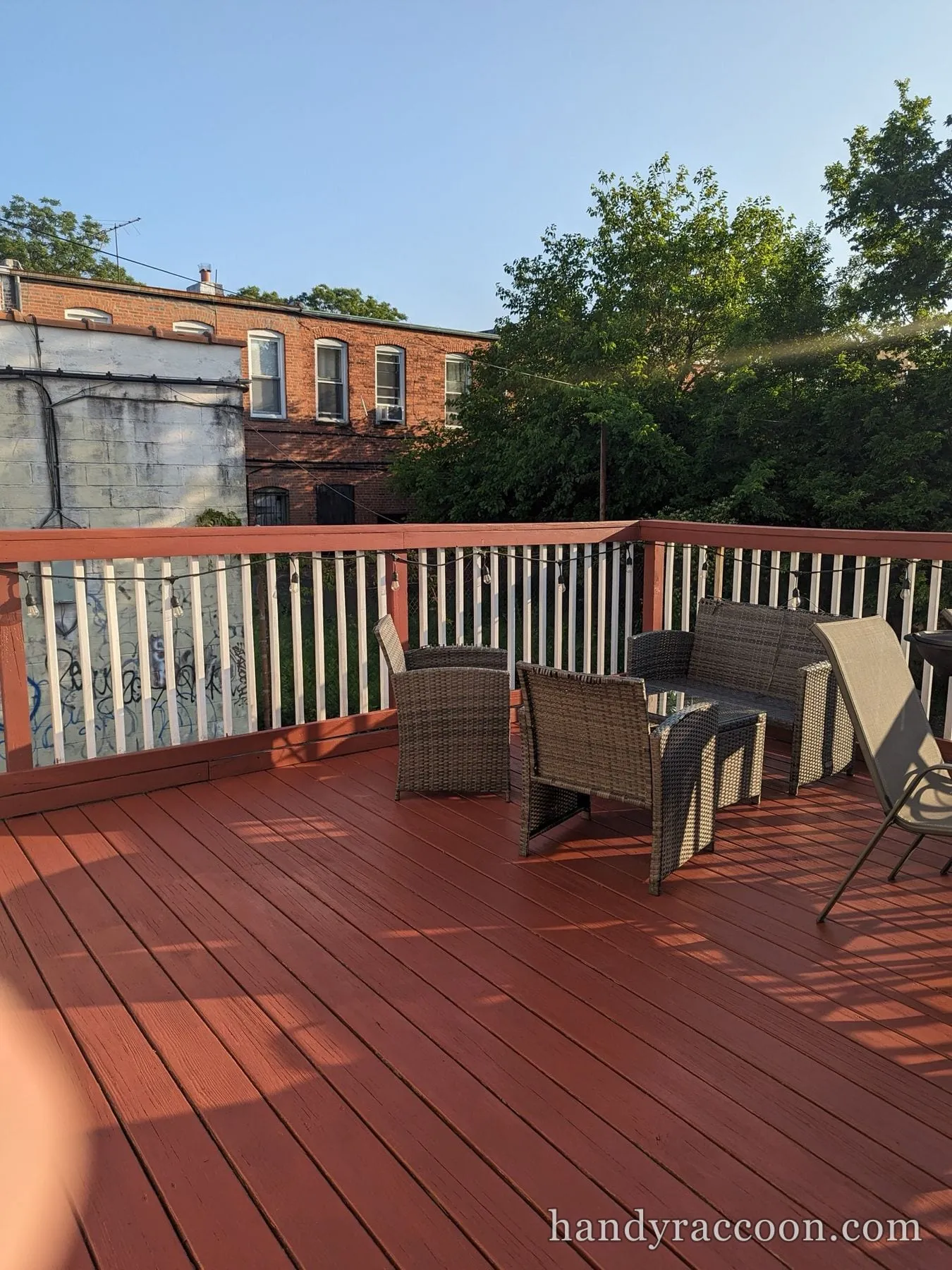 Maroon deck with wicker patio furniture in warm evening light, brick building behind.