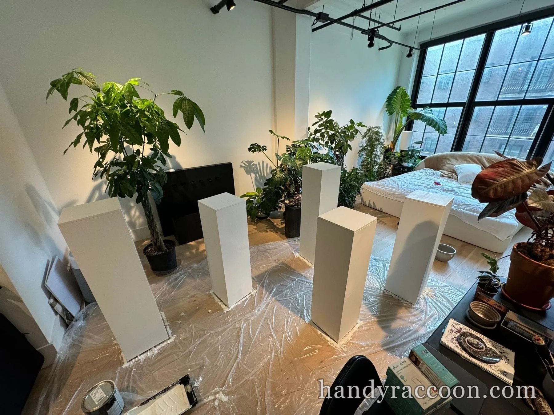 Several tall white painted pedestals standing on plastic drop cloth in a plant-filled loft.