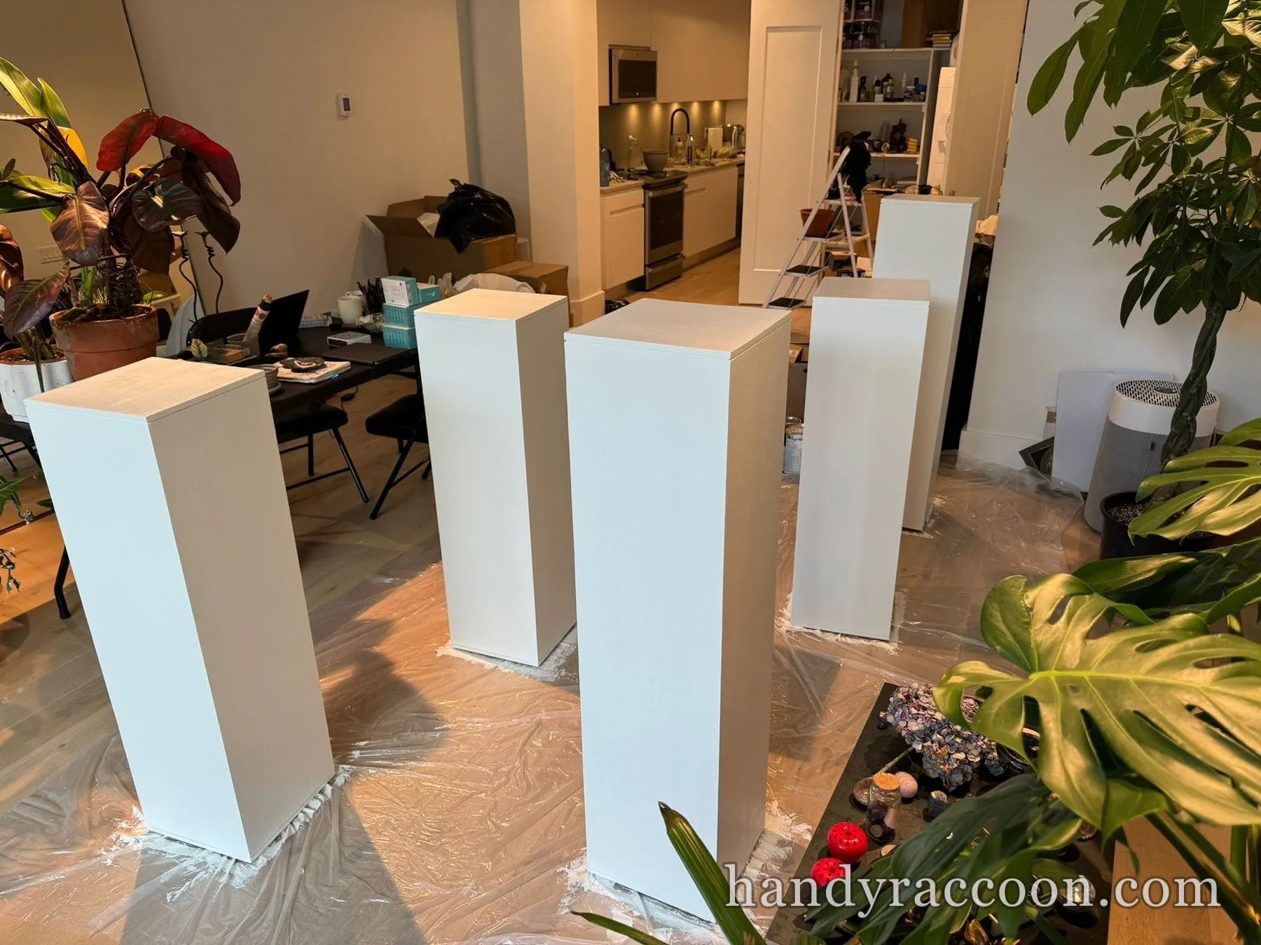 Four tall white rectangular pedestals standing upright on a drop cloth in a loft with plants.