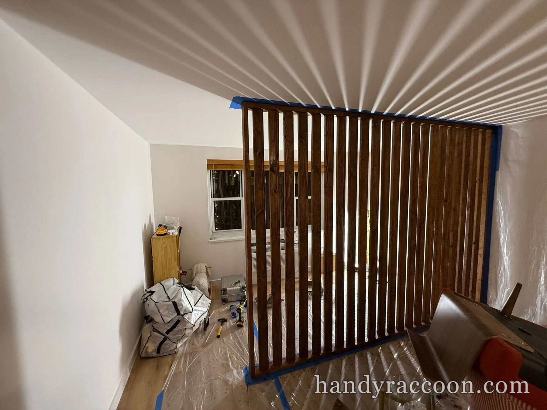 Vertical stained wood slat partition with sunlight casting stripes across the ceiling, drop cloth below.