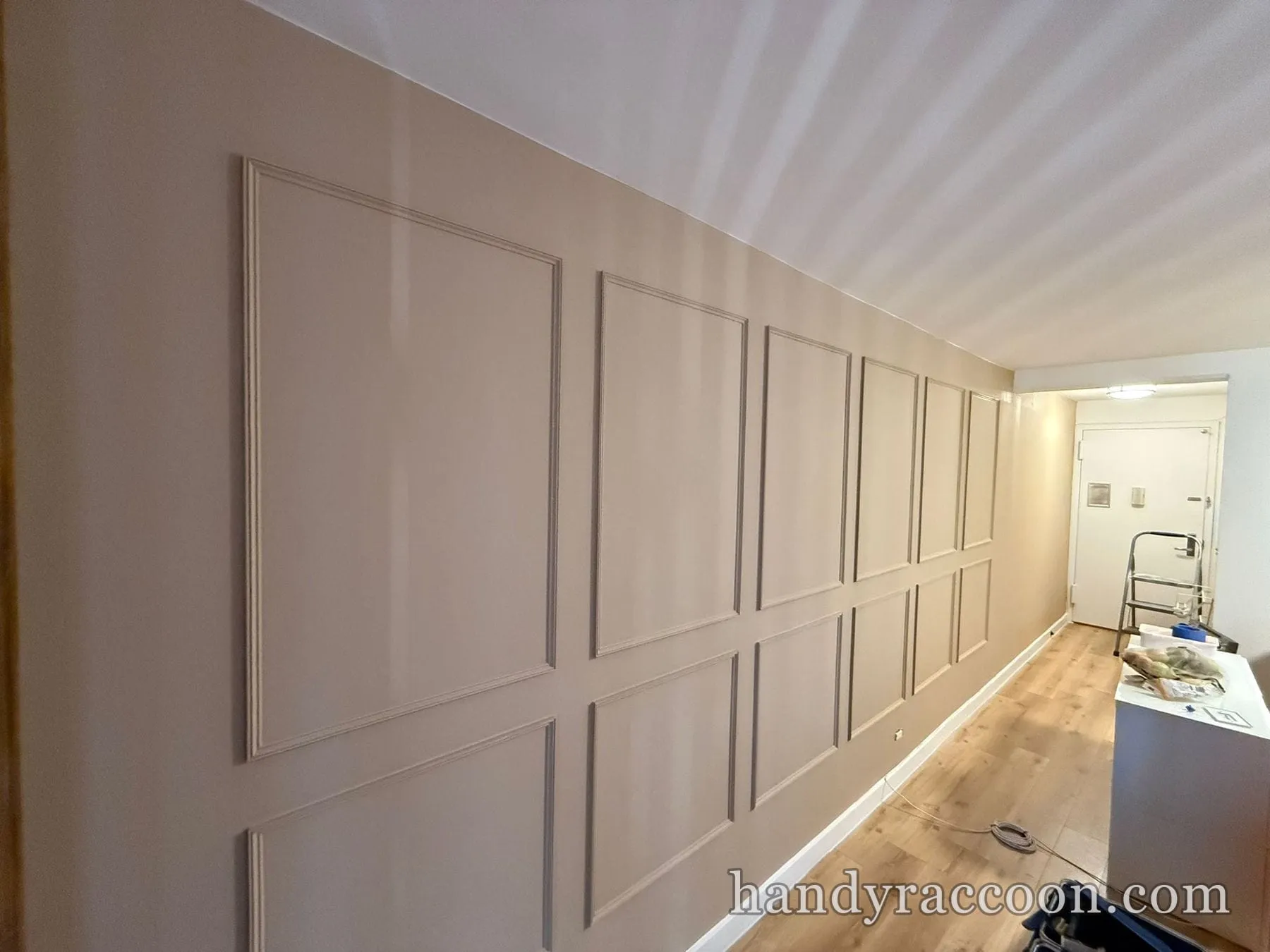 Beige hallway wall with evenly spaced rectangular panel molding leading to an entry door.