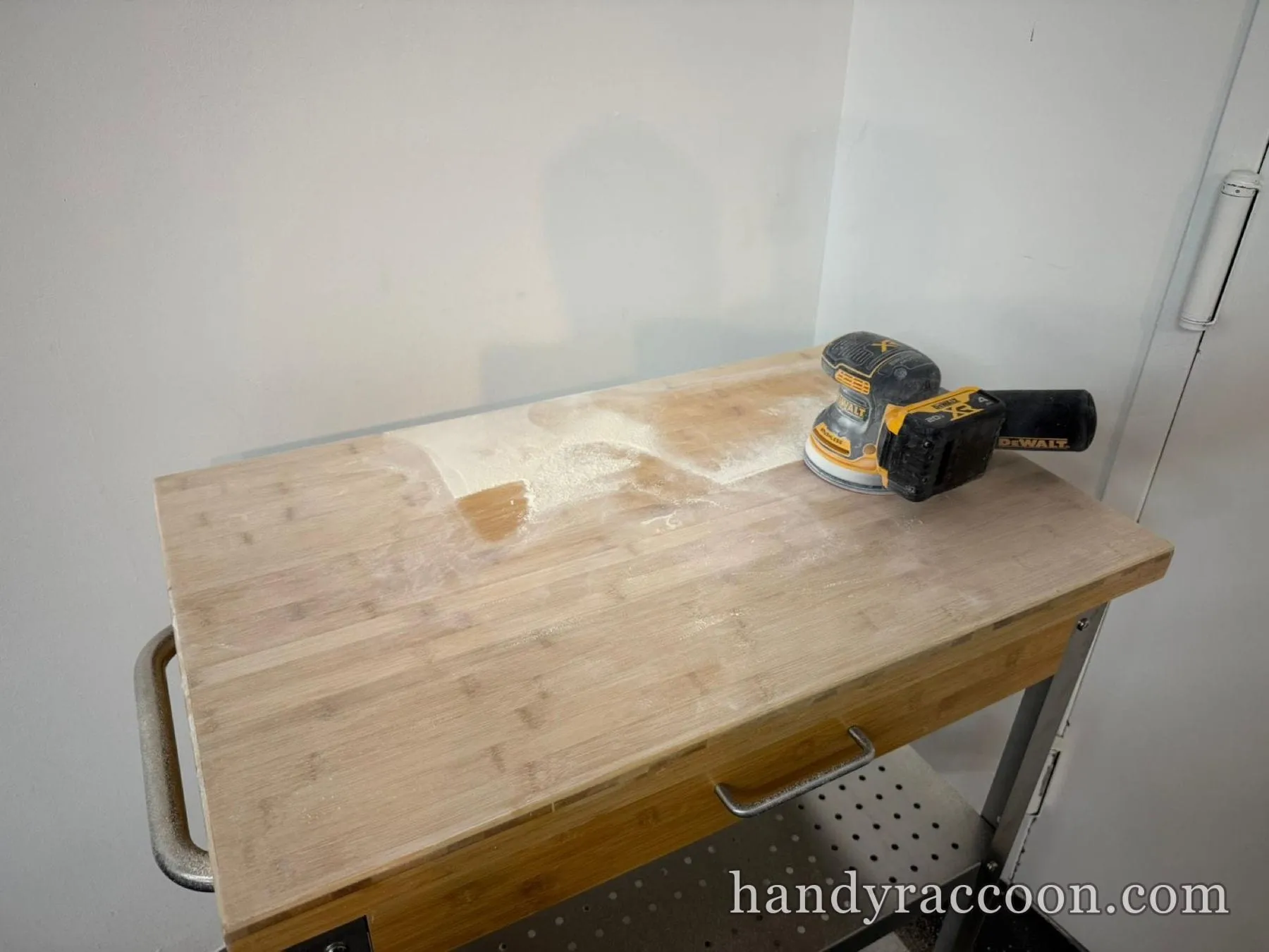 Wood butcher-block cart top with an orbital sander and sanding dust on top against a white wall.