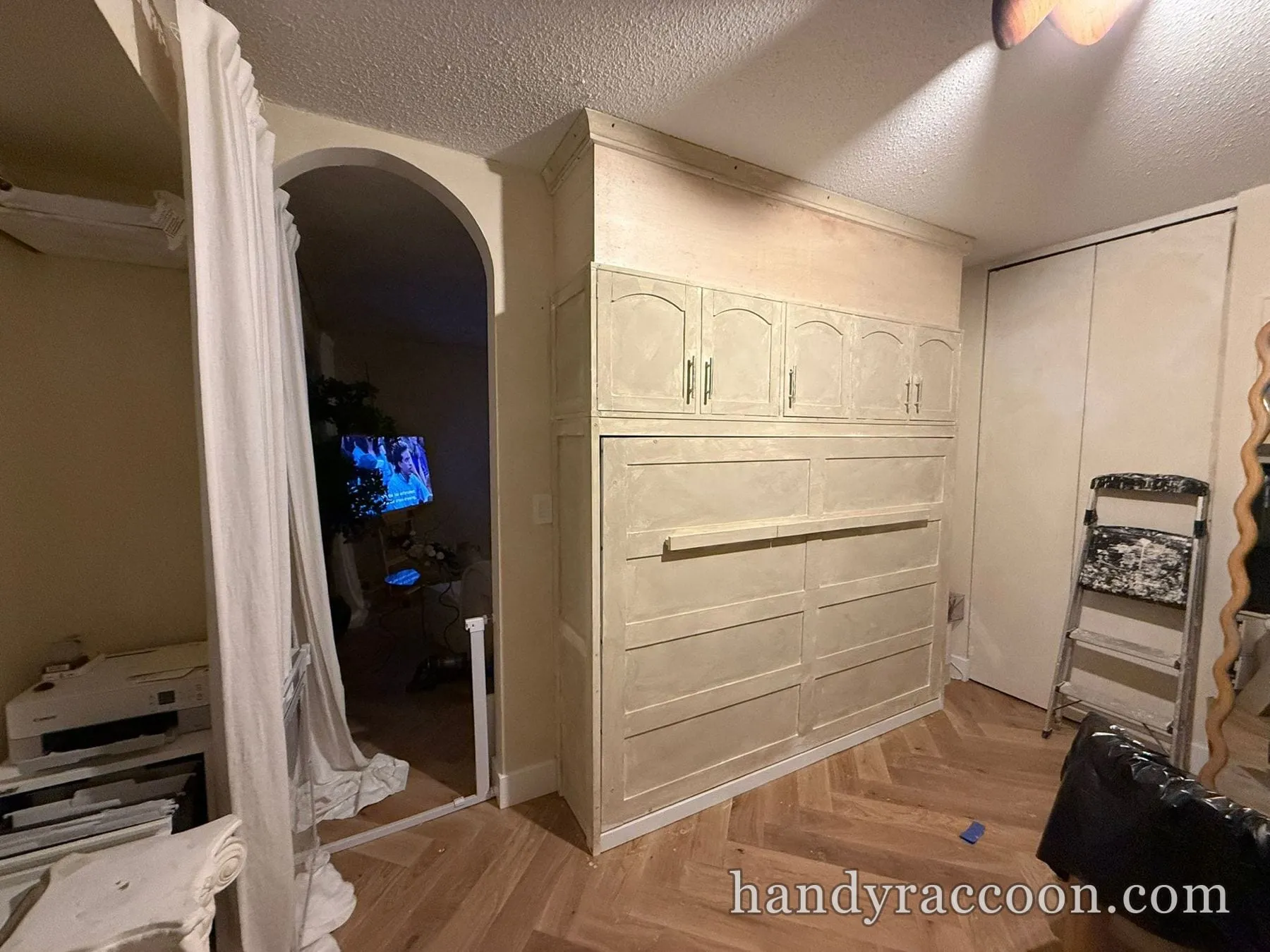 Large built-in cabinet and fold-down bed unit in raw primer against a bedroom wall with herringbone floor.