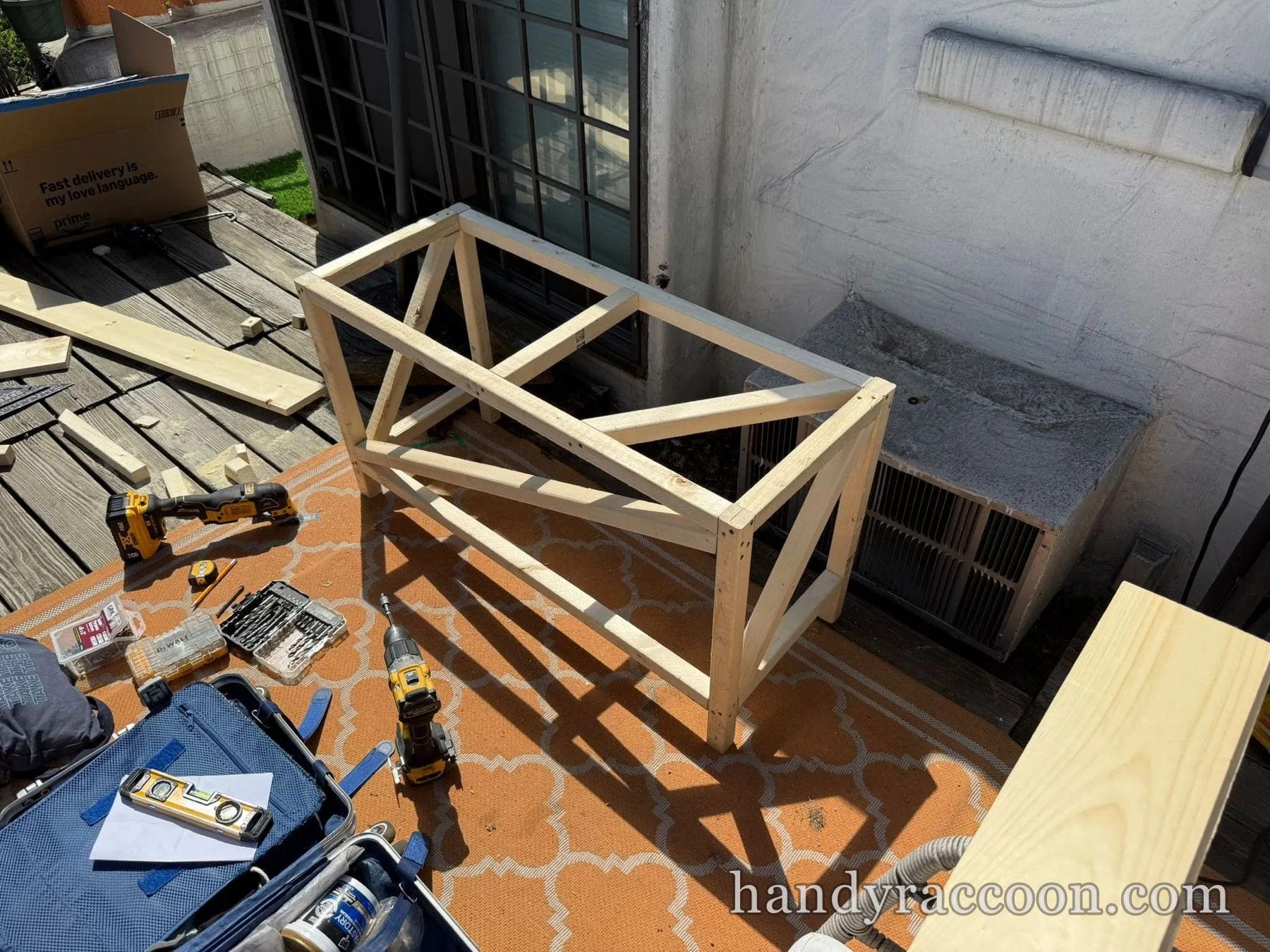 Partly built wooden X-frame table base on a patterned outdoor rug on a rooftop terrace with tools around.
