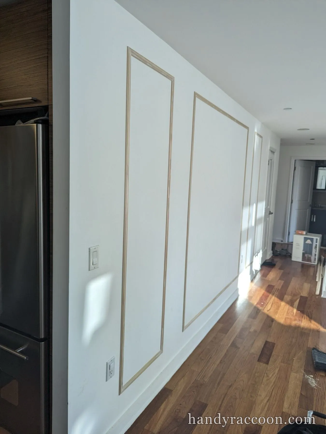 Unpainted wood picture-frame molding on a white wall beside a stainless refrigerator over hardwood.