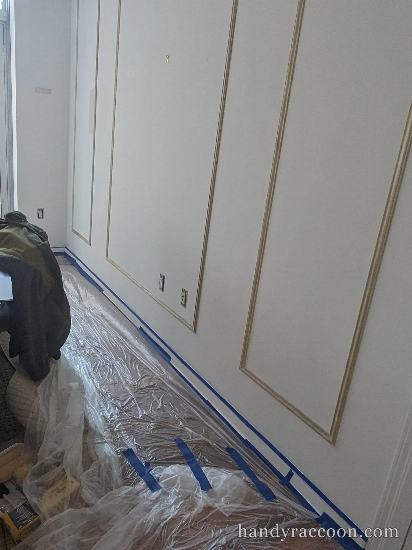White picture-frame wall molding with blue tape and floor sheeting during painting prep.