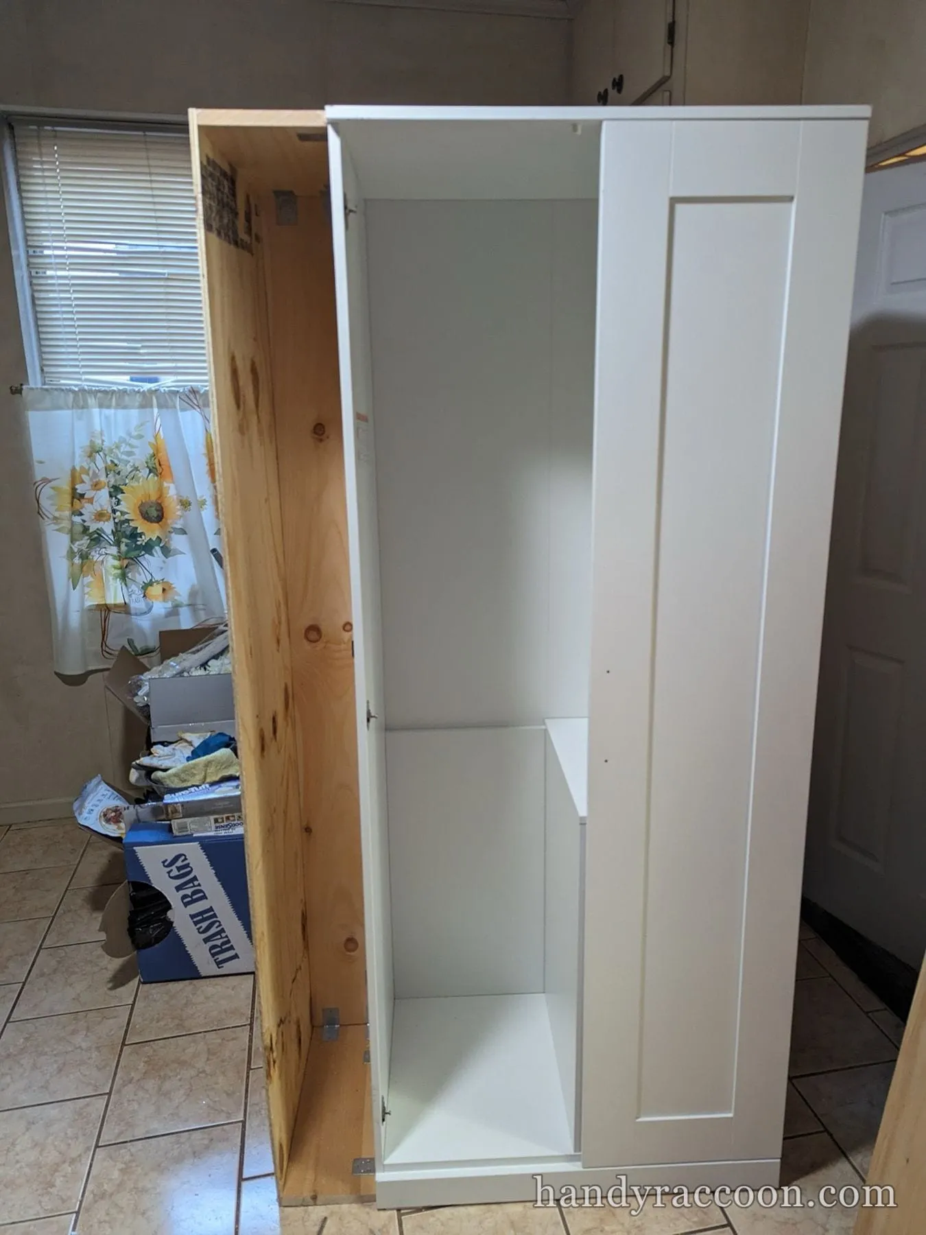 A tall white cabinet with a plywood side and open door standing on a tile kitchen floor.