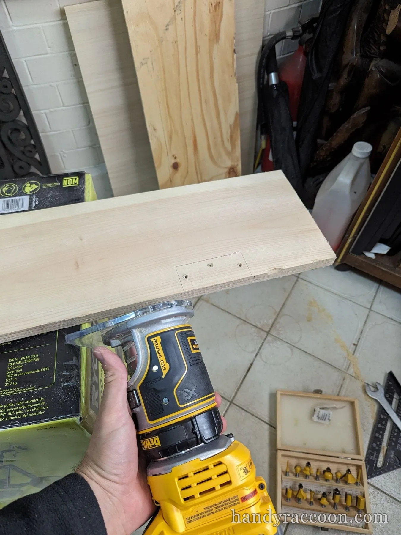 A hand guides a yellow cordless router cutting a shallow recess in a pine board in a garage.