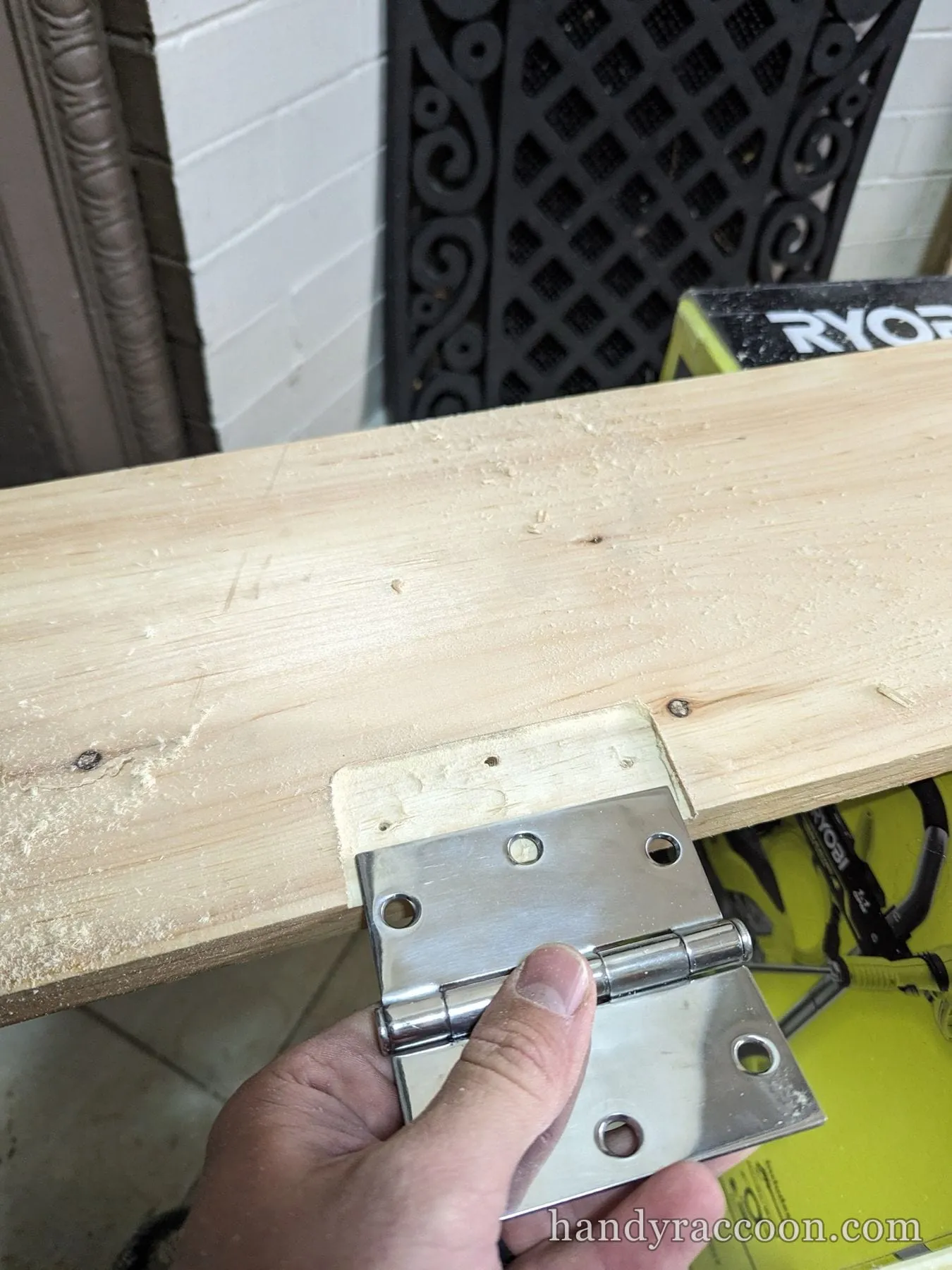 A hand holds a chrome butt hinge against a mortise cut into the edge of a pine board.