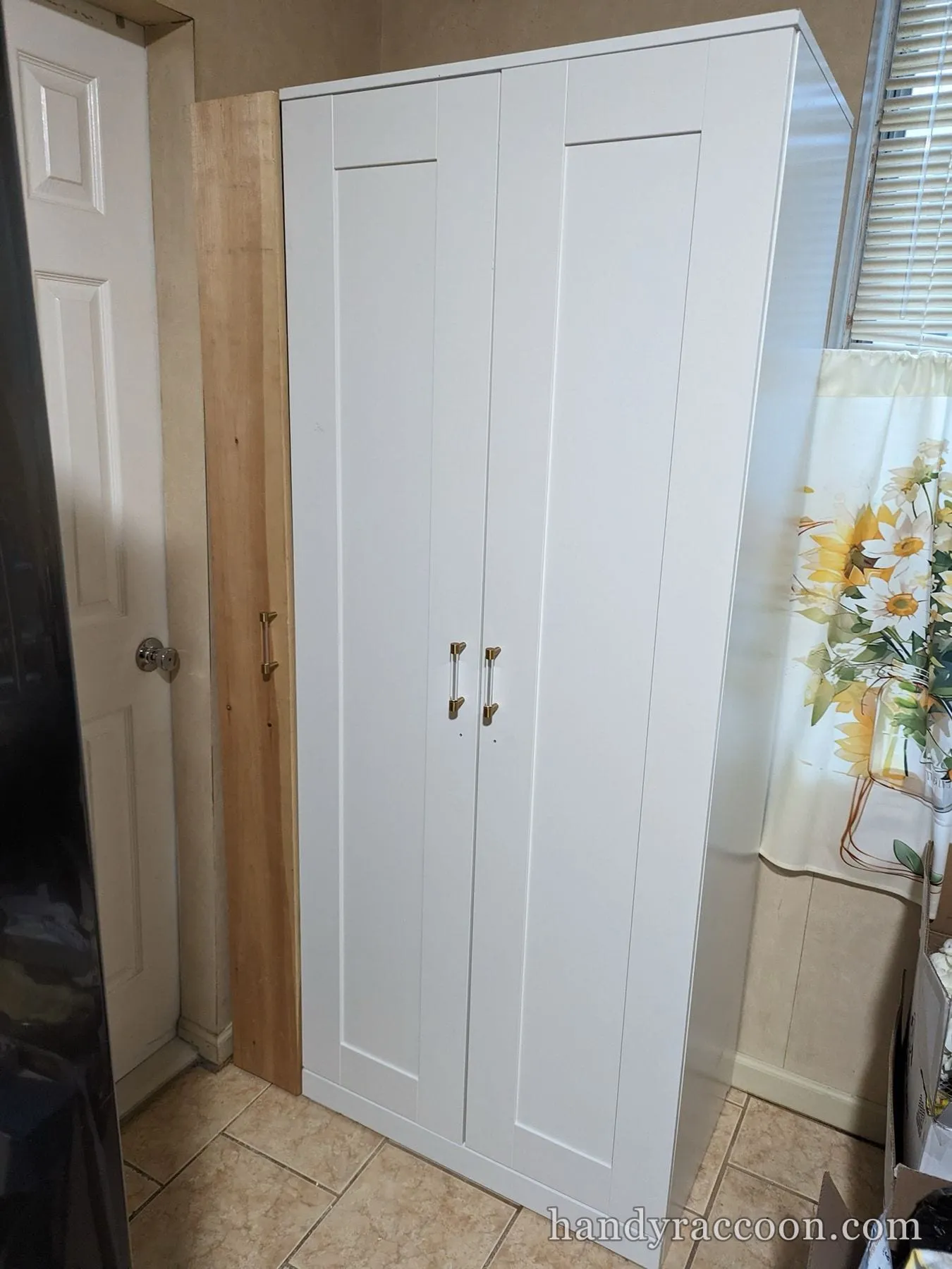 A tall white two-door shaker cabinet with glass and gold handles standing in a room corner.